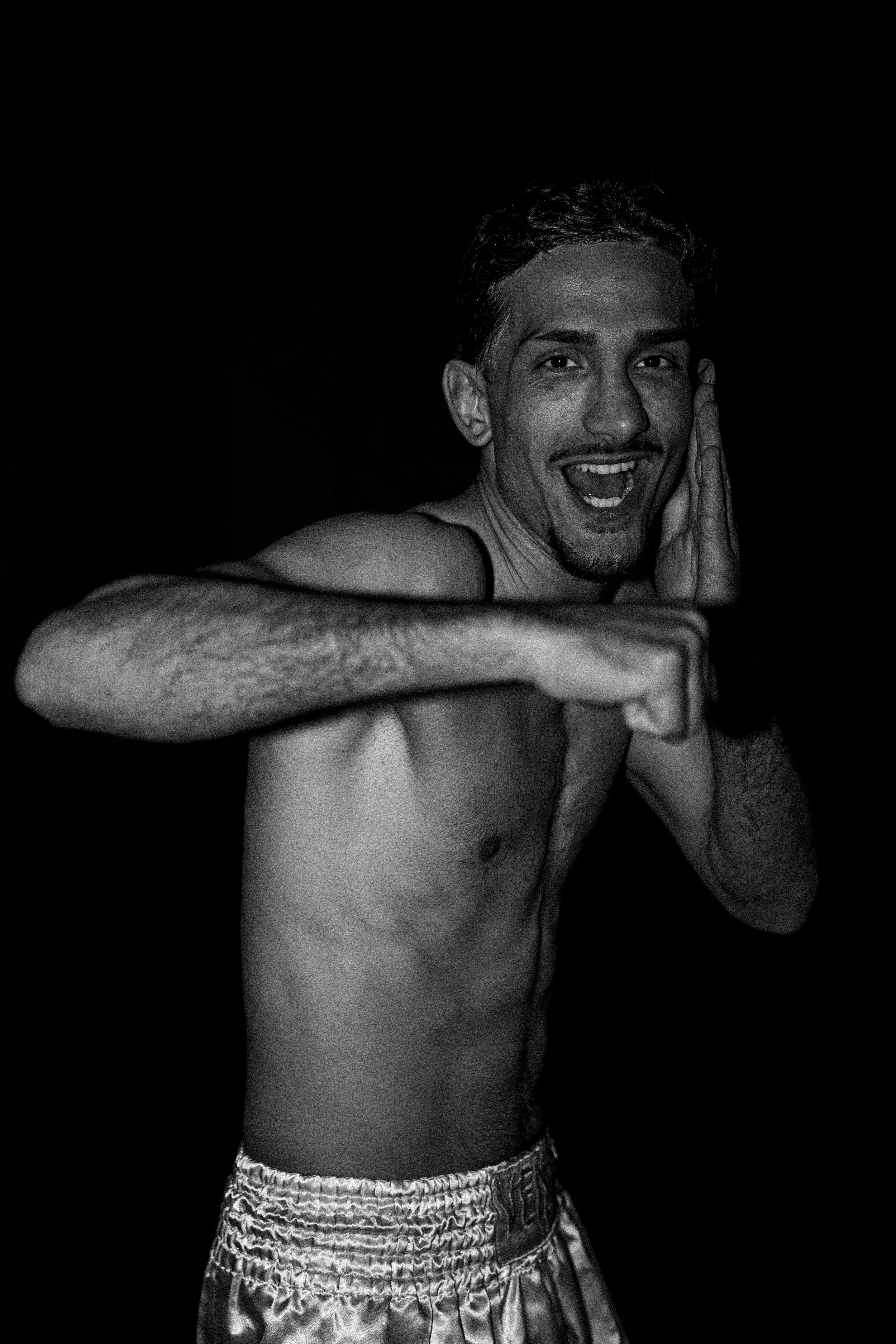 A shirtless man with a goatee, smiling with his mouth open, holding his hand near his face, and pointing his finger towards the camera. He is wearing shorts, set against a dark background.