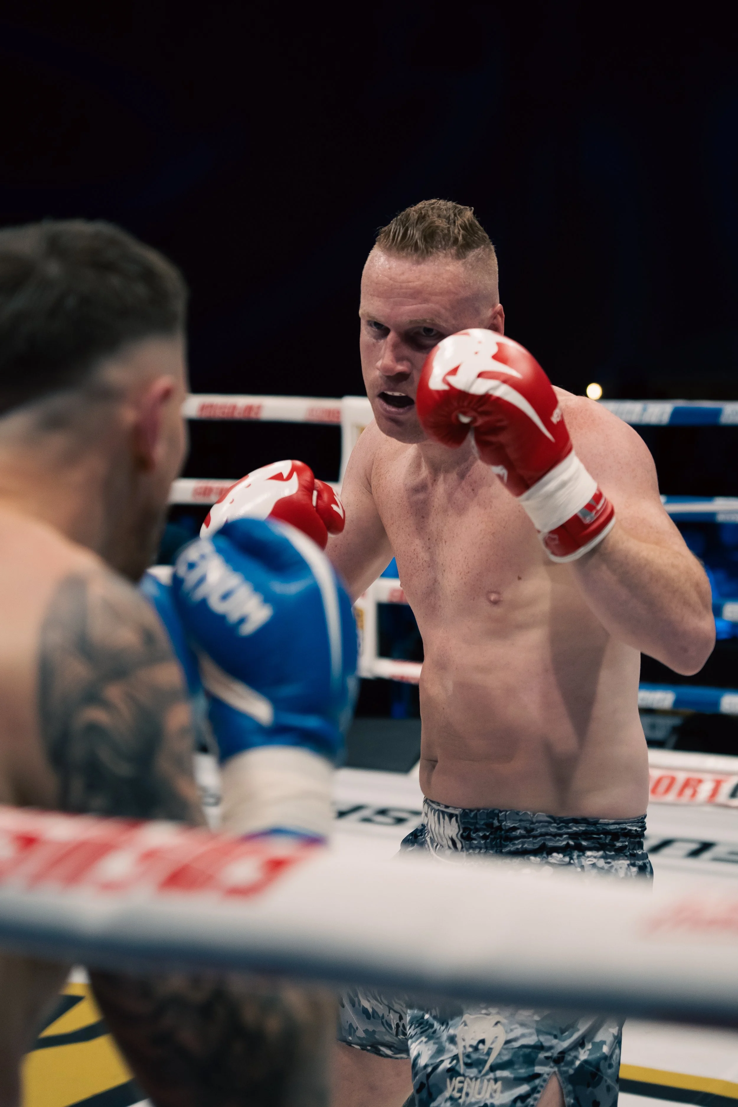 Two male boxers in a boxing ring engaged in a match, one shirtless wearing red gloves and patterned shorts, the other partially visible with blue gloves, fighting intensely.