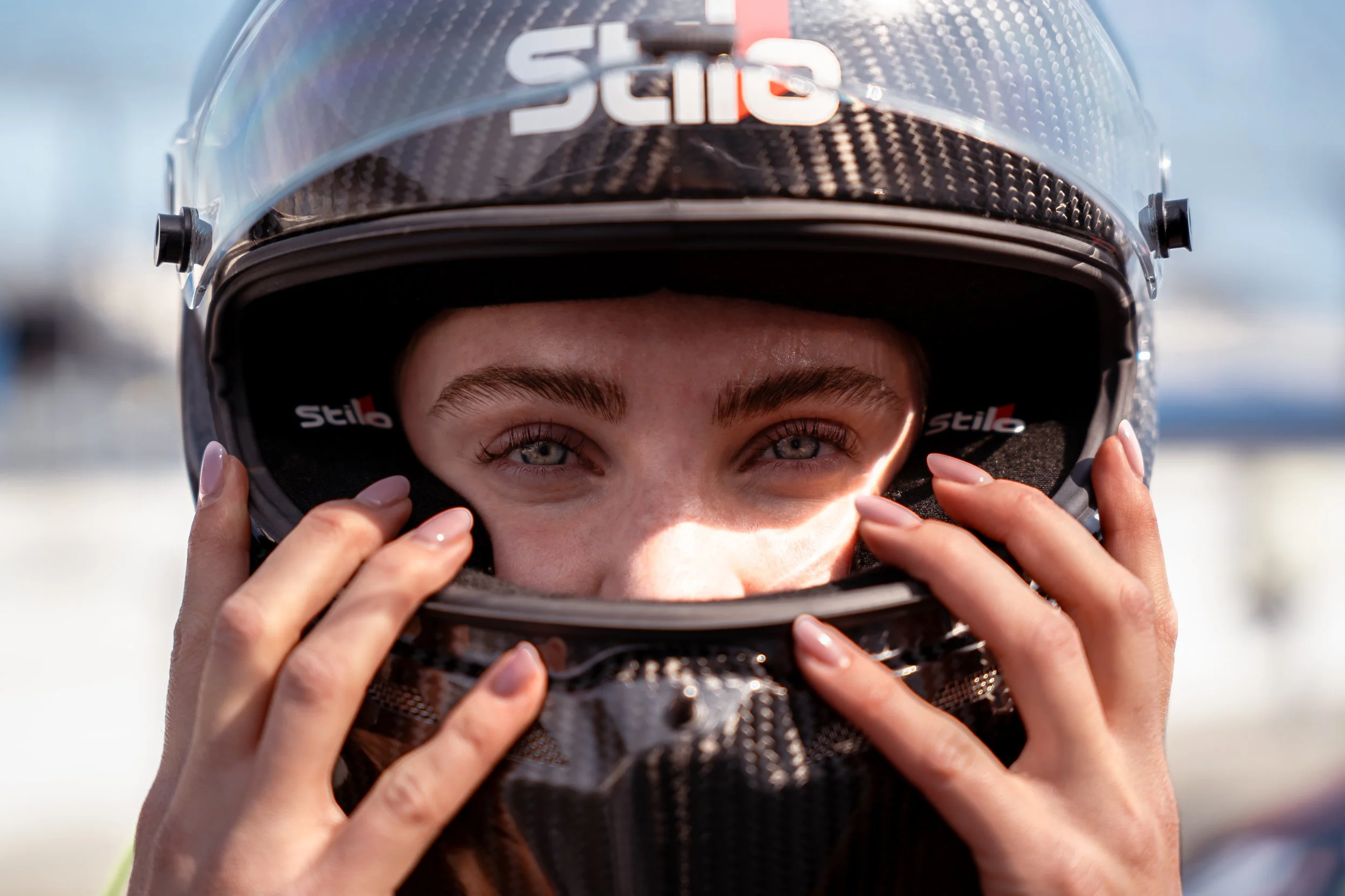 Close-up of a person wearing a motorcycle helmet, adjusting it with both hands. The helmet is black with a carbon fiber pattern and a clear visor. The person's eyes are visible, looking directly at the camera.