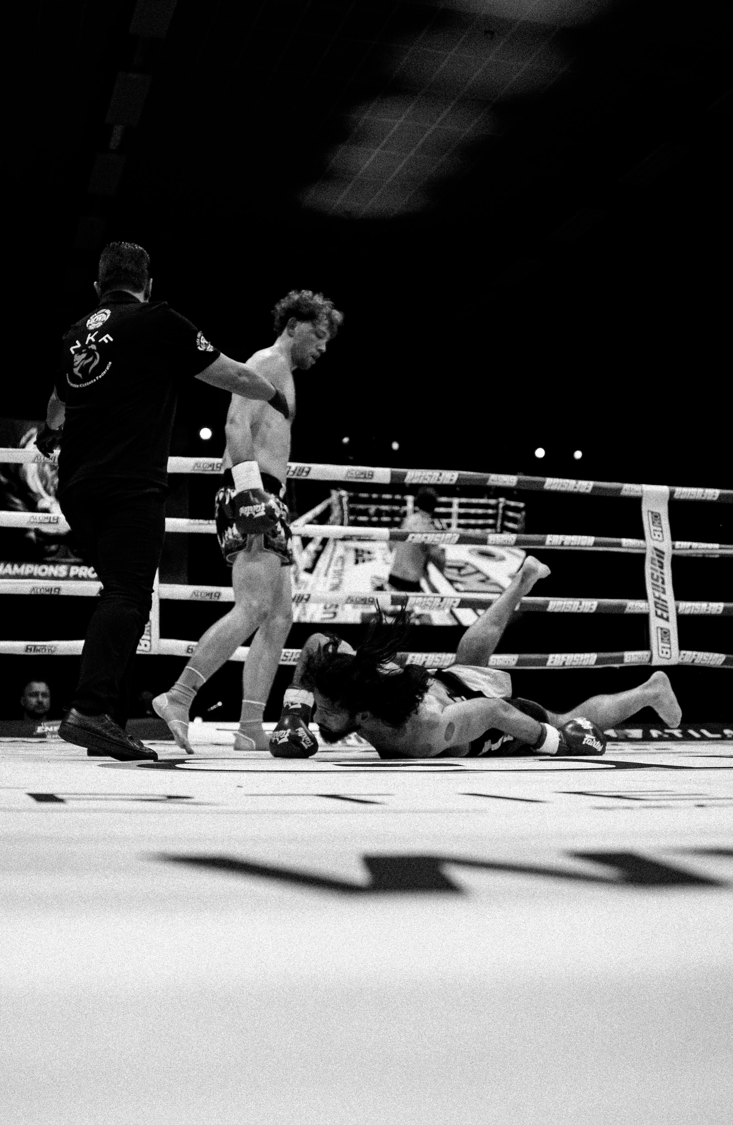 Black and white photo of two male Muay Thai fighters, one on the ground and another standing, inside a boxing ring with a referee nearby, during a match.