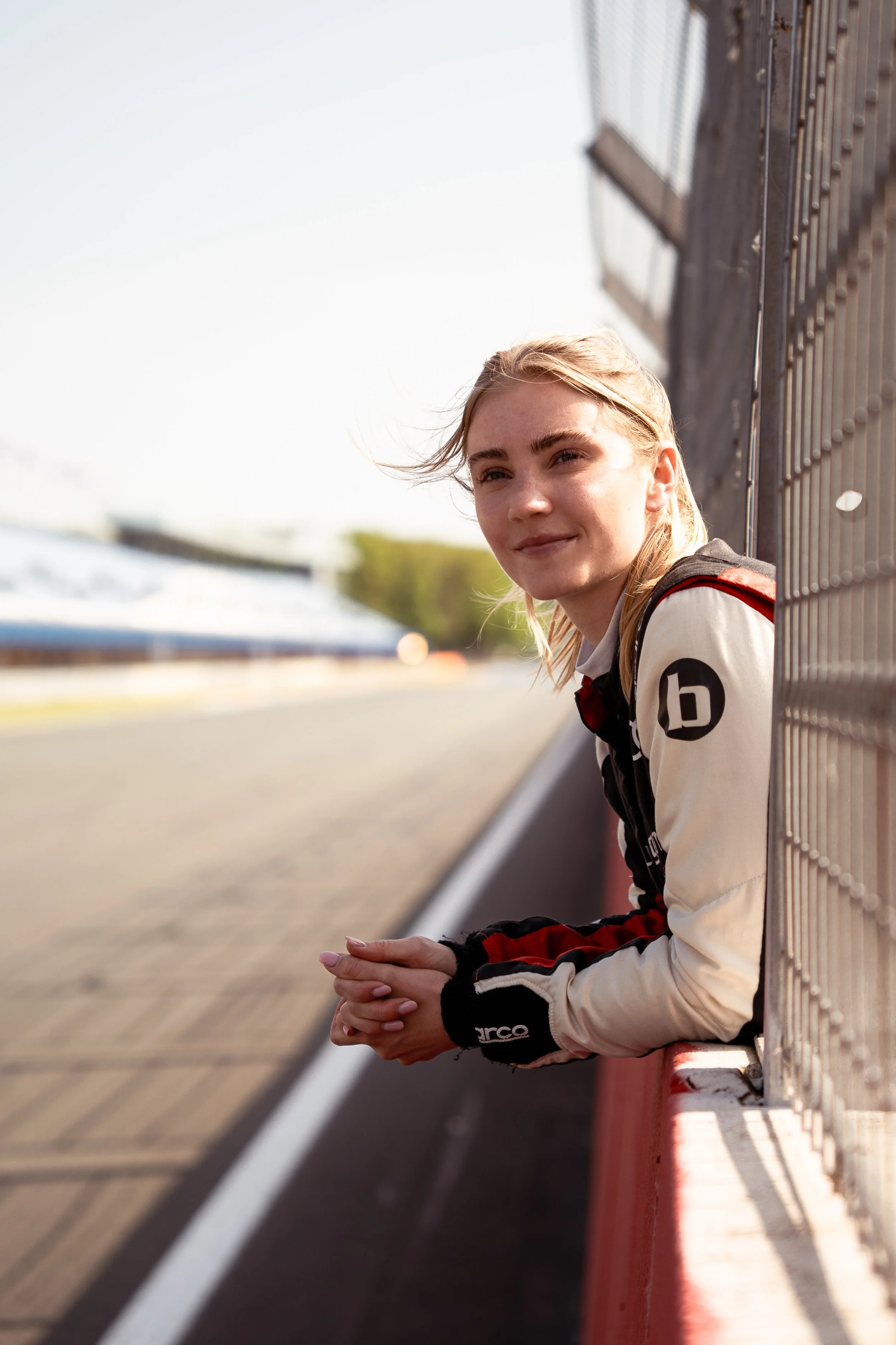 A young woman with blonde hair smiling, wearing a racing jacket, sitting on a racing track barrier.