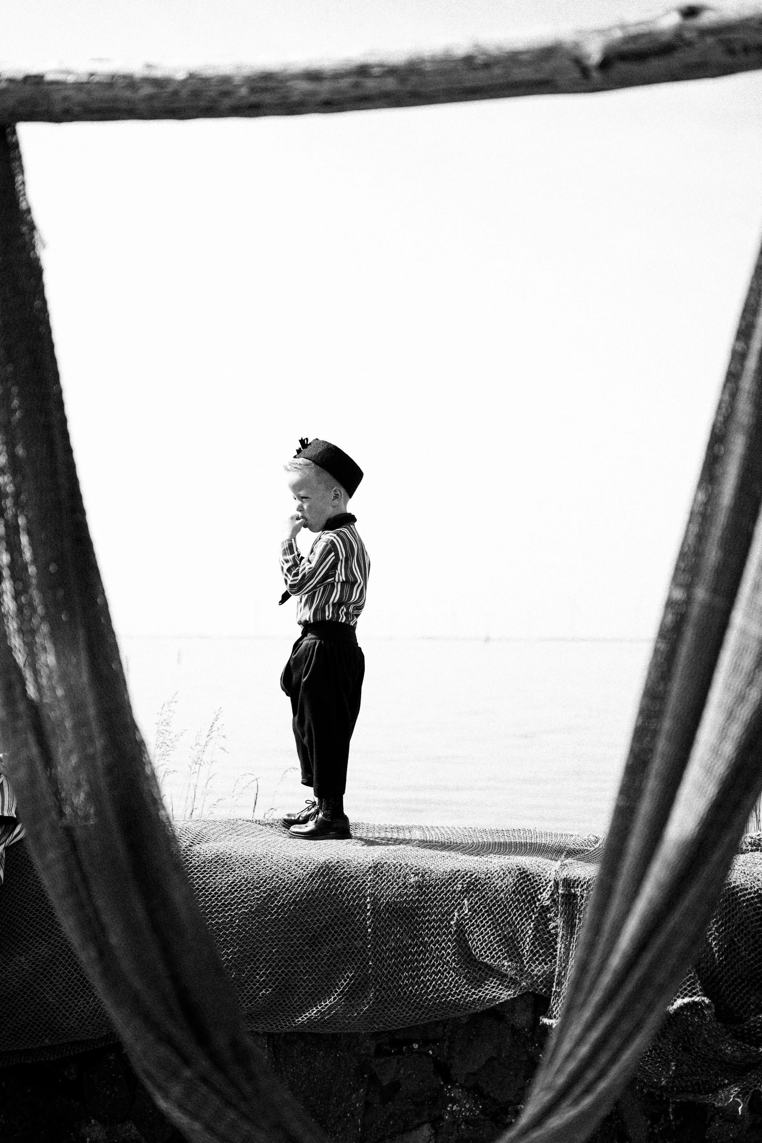 A young boy dressed in striped shirt and dark pants, standing on a patterned surface outdoors, framed by the slats of a wooden structure.