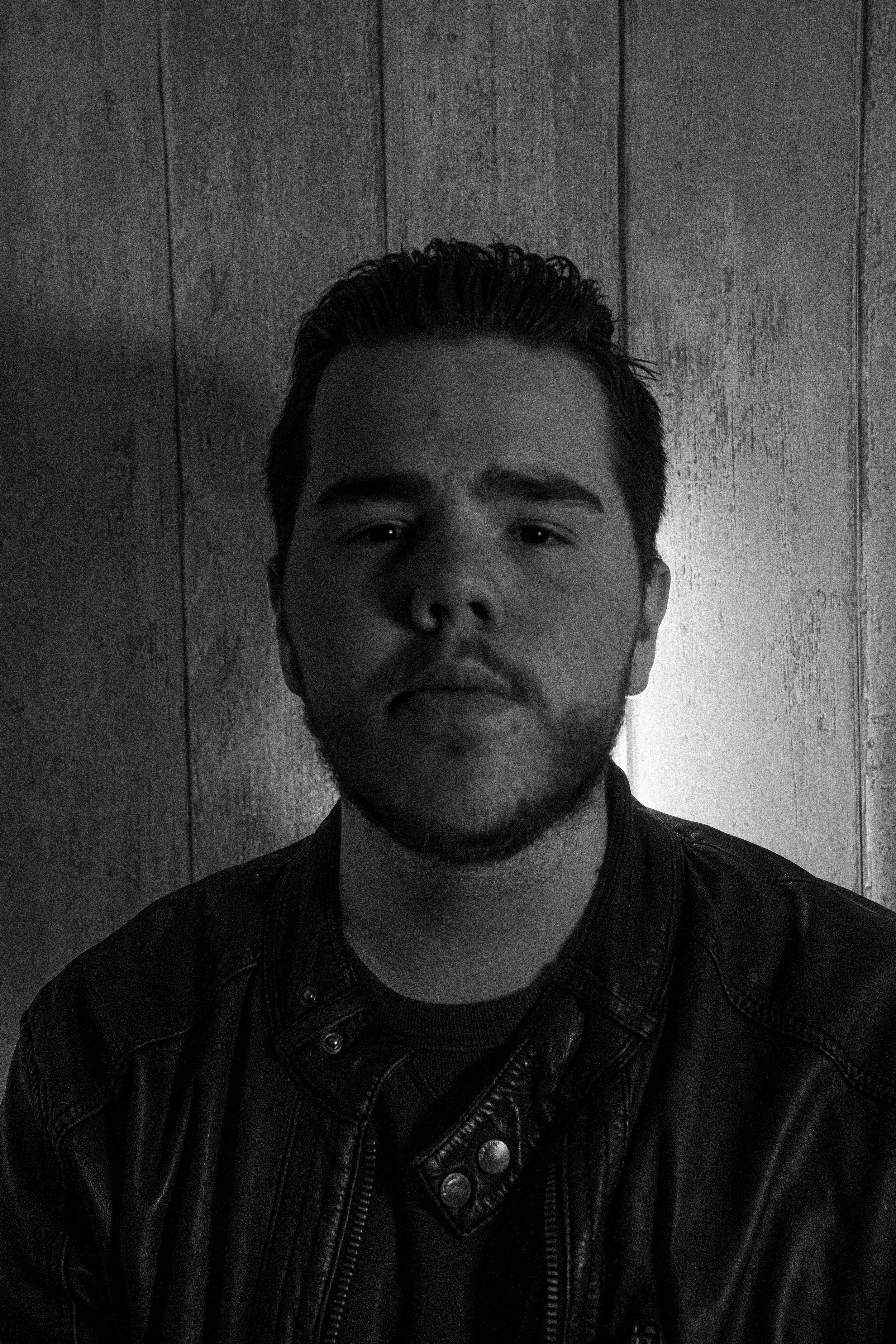 Young man with dark hair and facial hair wearing a leather jacket, looking directly at the camera, standing against a wooden wall background.
