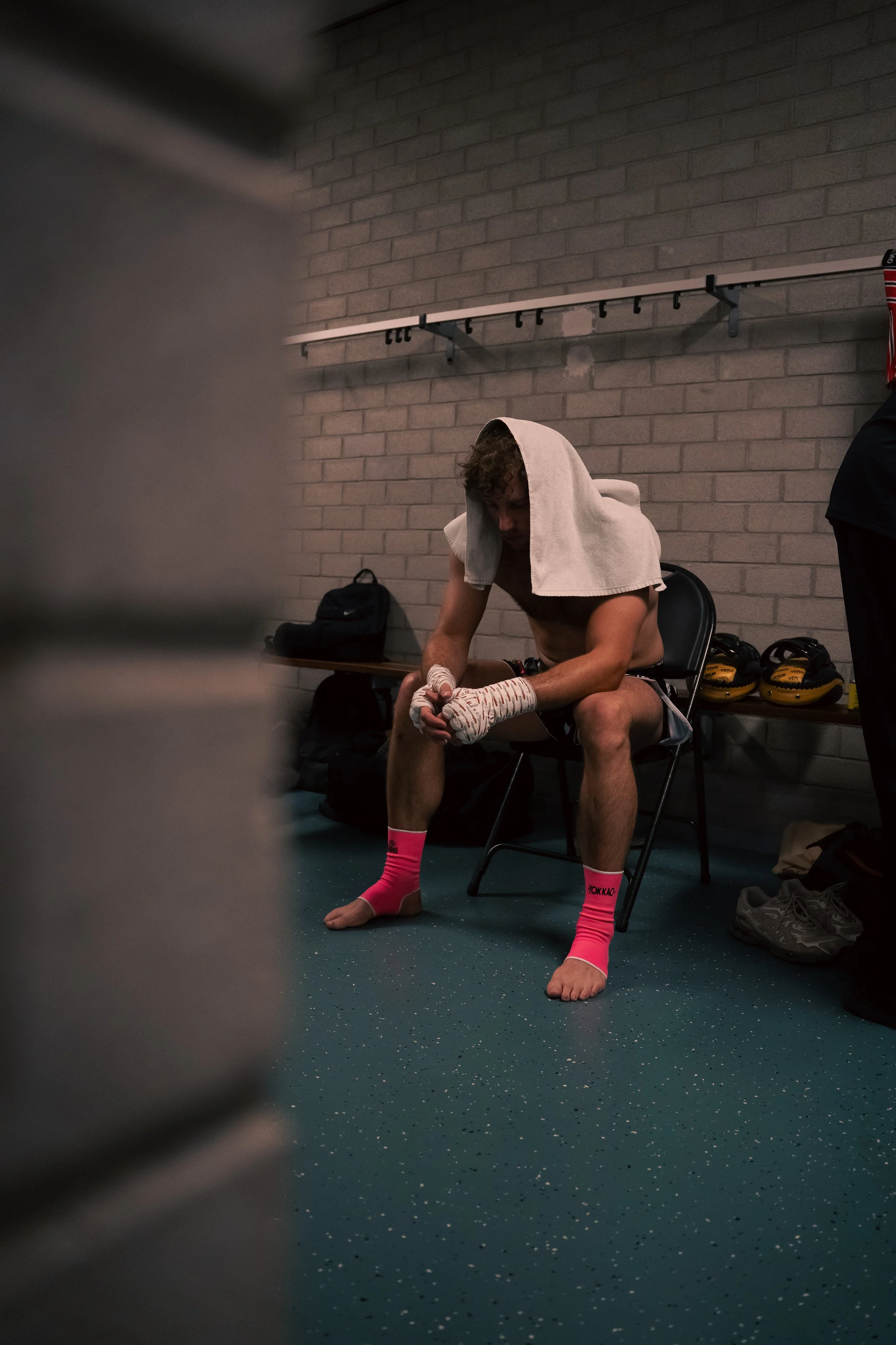 A shirtless male boxer with a towel over his head, sitting on a chair in a locker room, tying his boxing hand wraps. He is wearing pink boxing socks and has boxing gloves on the floor nearby. The background features a brick wall and a row of lockers.