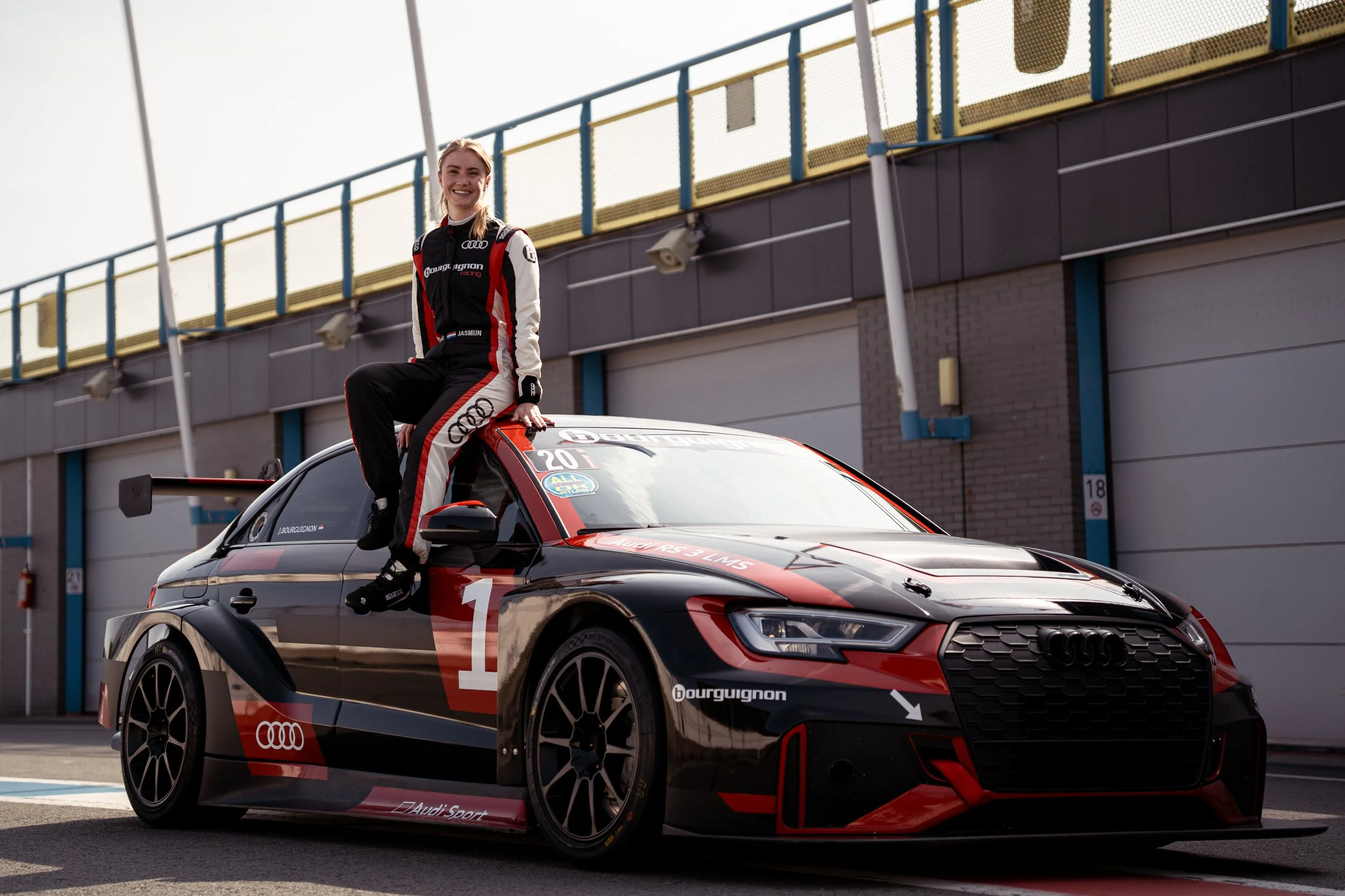 Female race car driver in racing suit sitting on the roof of a black and red Audi race car at a race track, smiling.