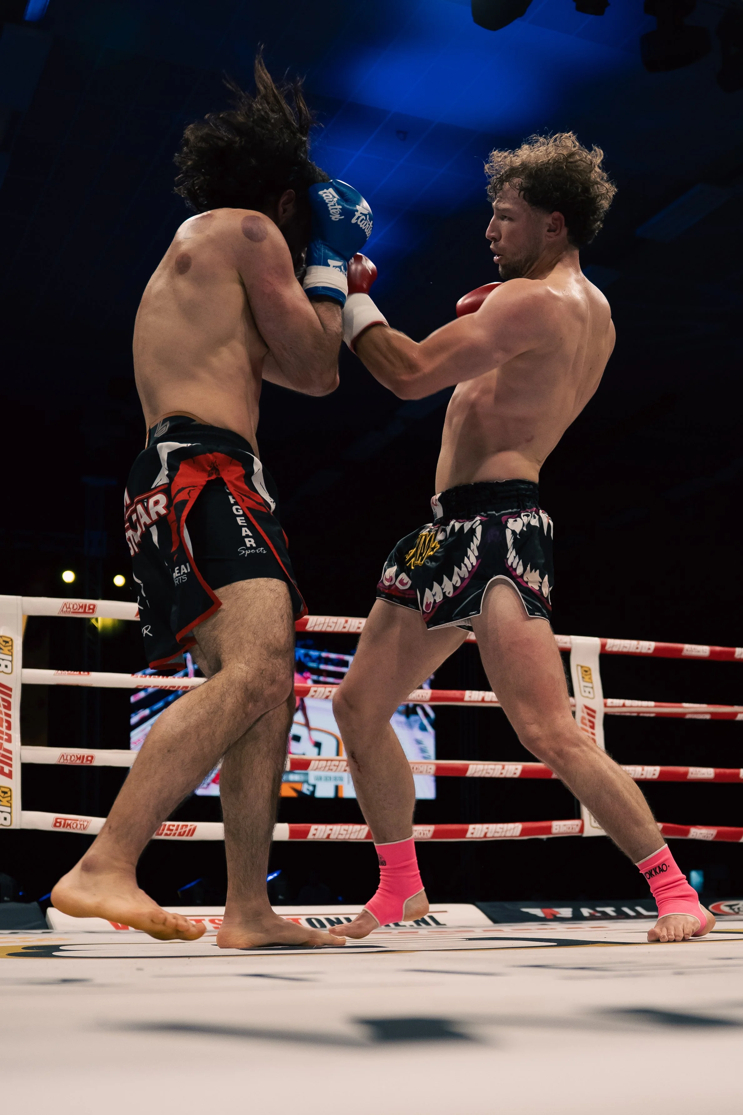Two male fighters in boxing gloves exchanging punches inside a boxing ring during a match, with a dark background and bright lights overhead.