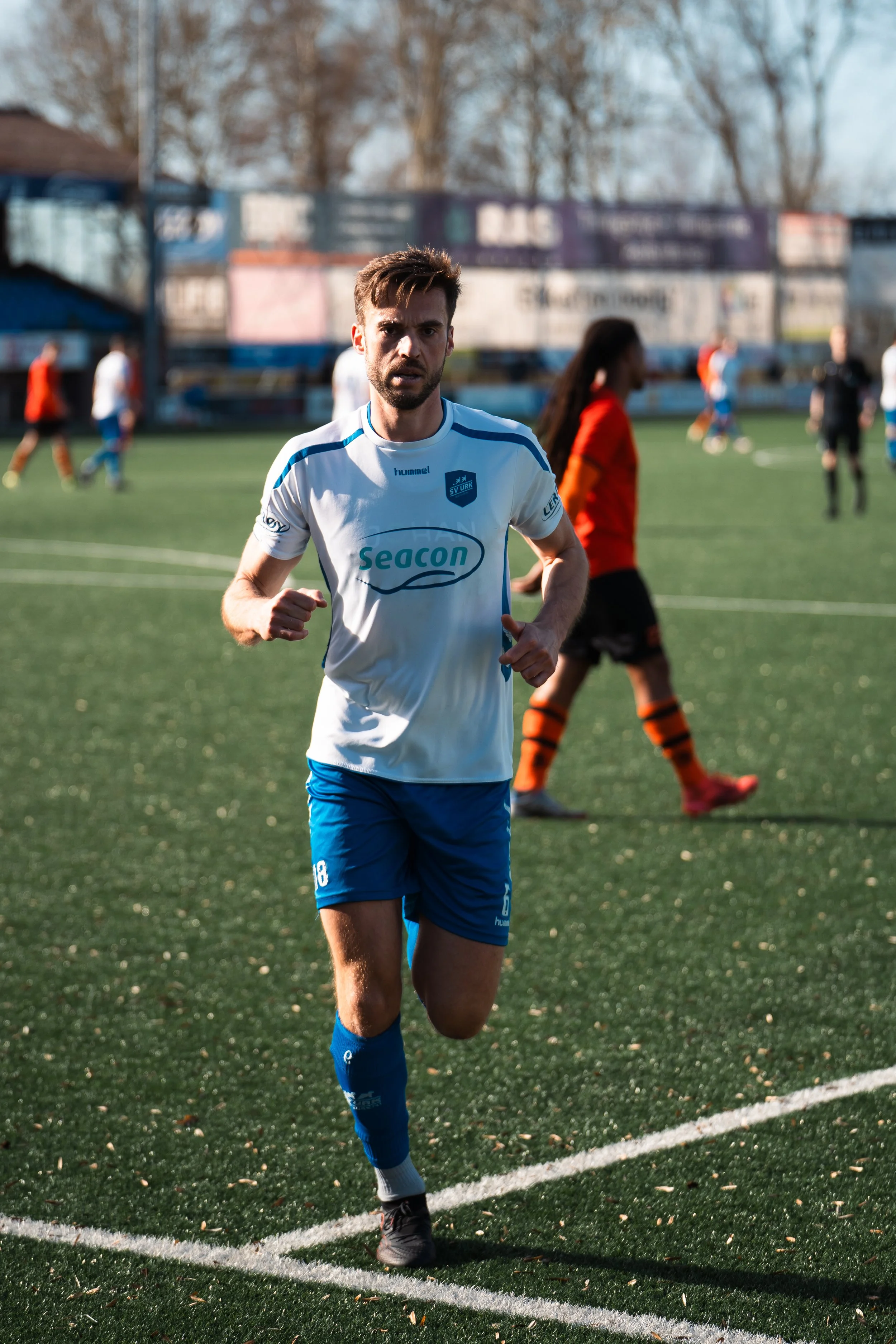 A male soccer player in a white and blue uniform jogging on a soccer field with other players and a referee in the background.