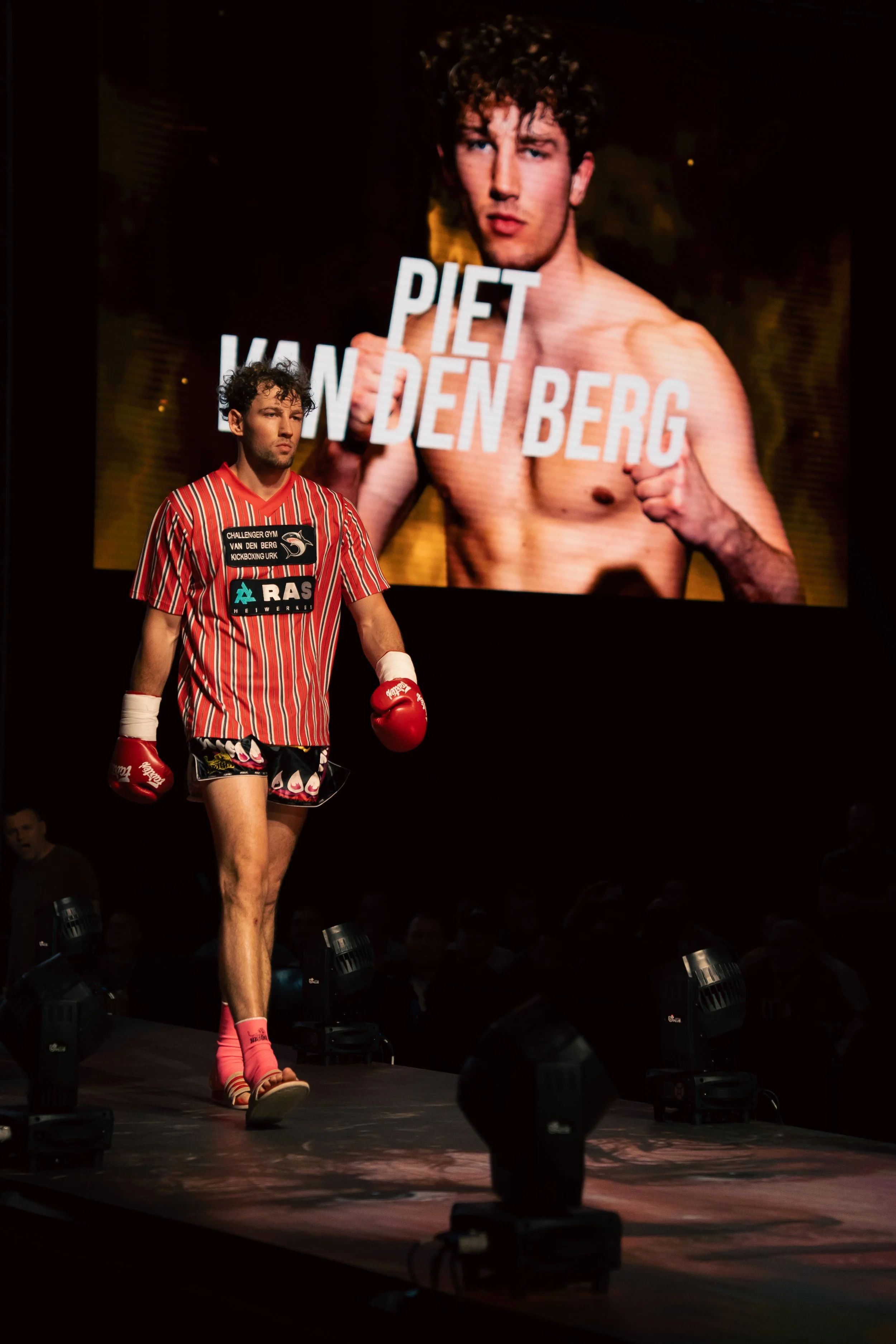A male athlete with curly hair in a red and black striped martial arts uniform, wearing red gloves and pink socks, walking on stage with a large screen behind him displaying a shirtless man with curly hair, holding boxing gloves, and text reading 'PI