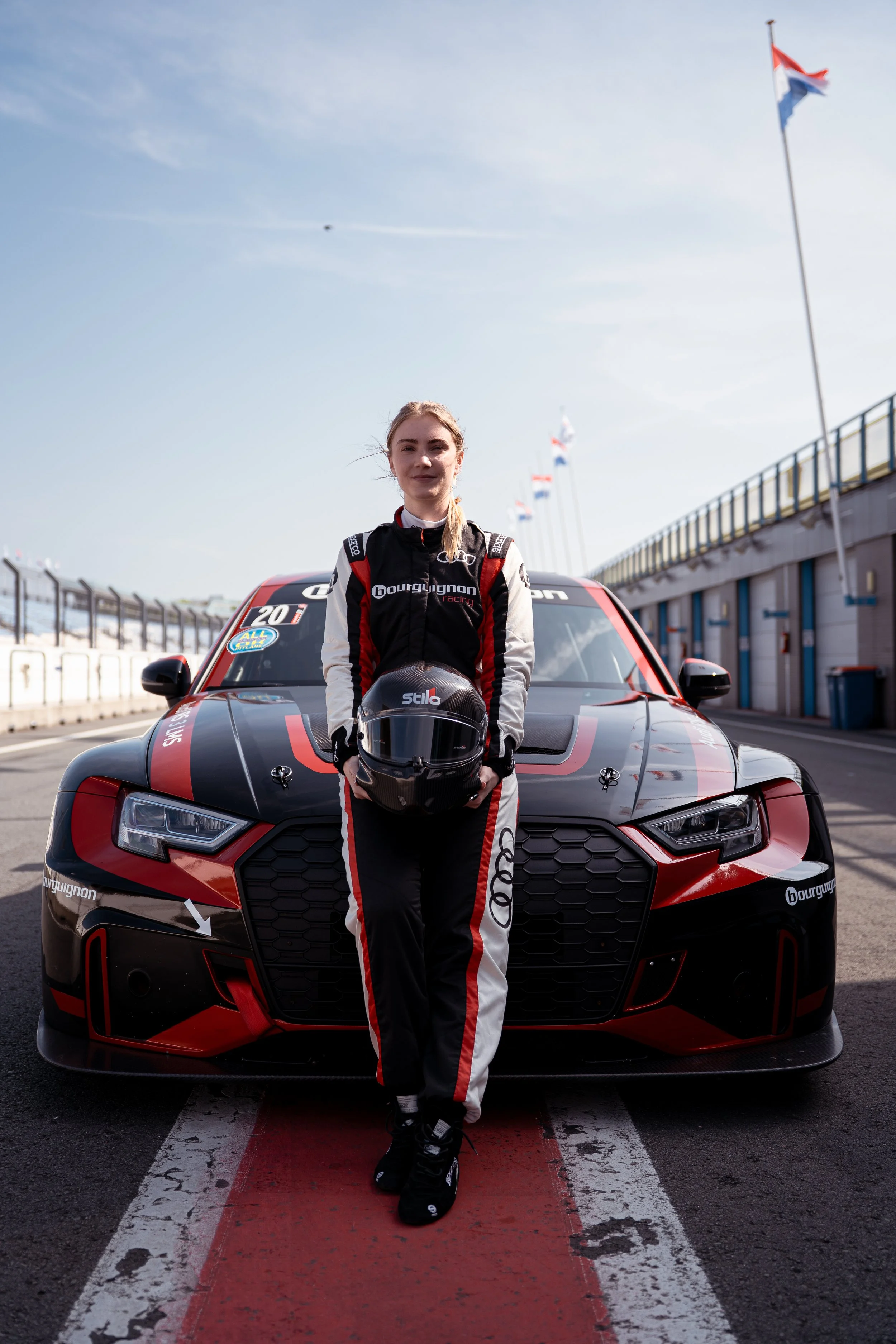 A woman in racing suit standing in front of a black and red race car on a racetrack with flags in the background.