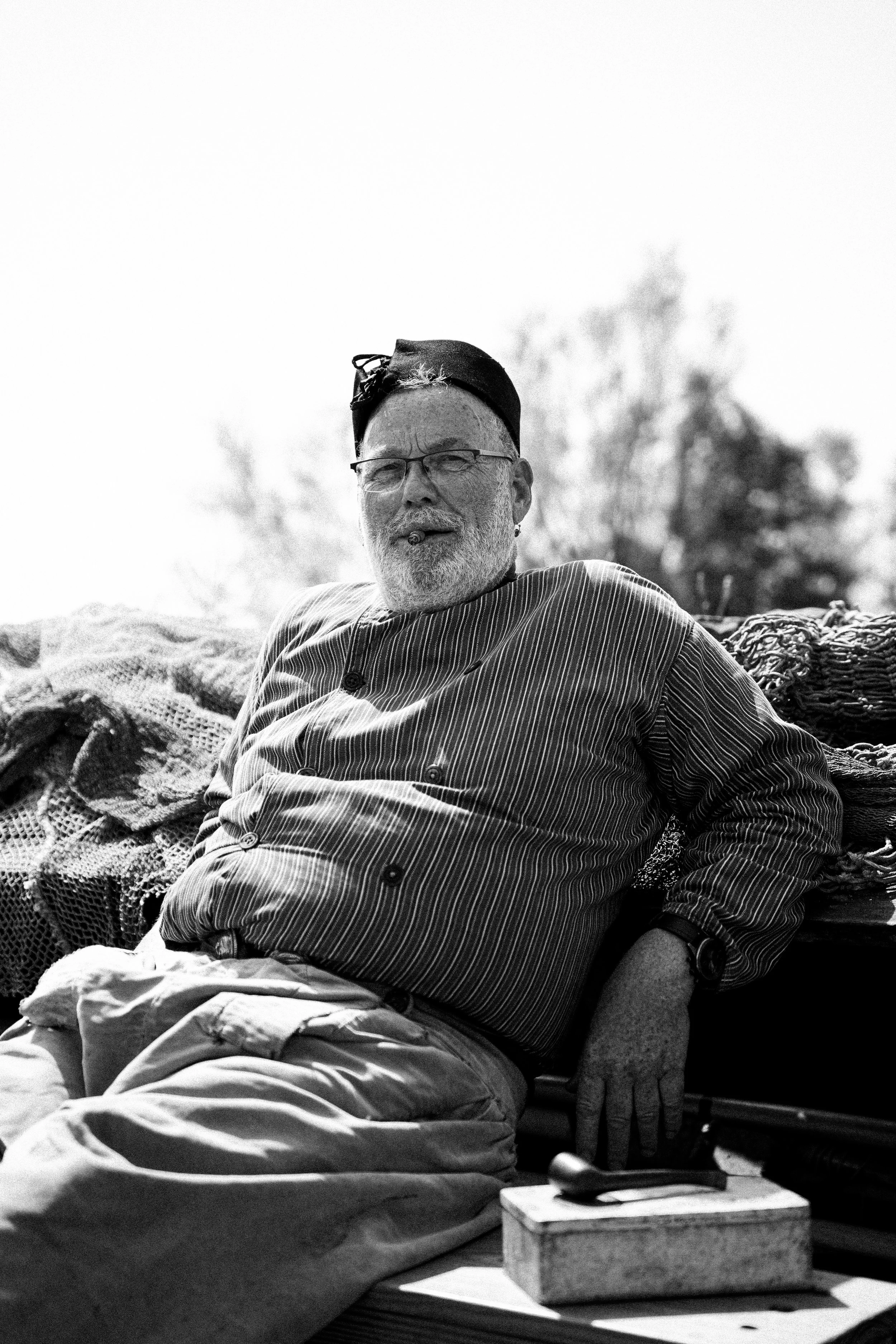 A black and white photo of an older man with glasses, a beard, and a cigarette in his mouth, wearing a hat and striped shirt, sitting outdoors on a bench with trees in the background.