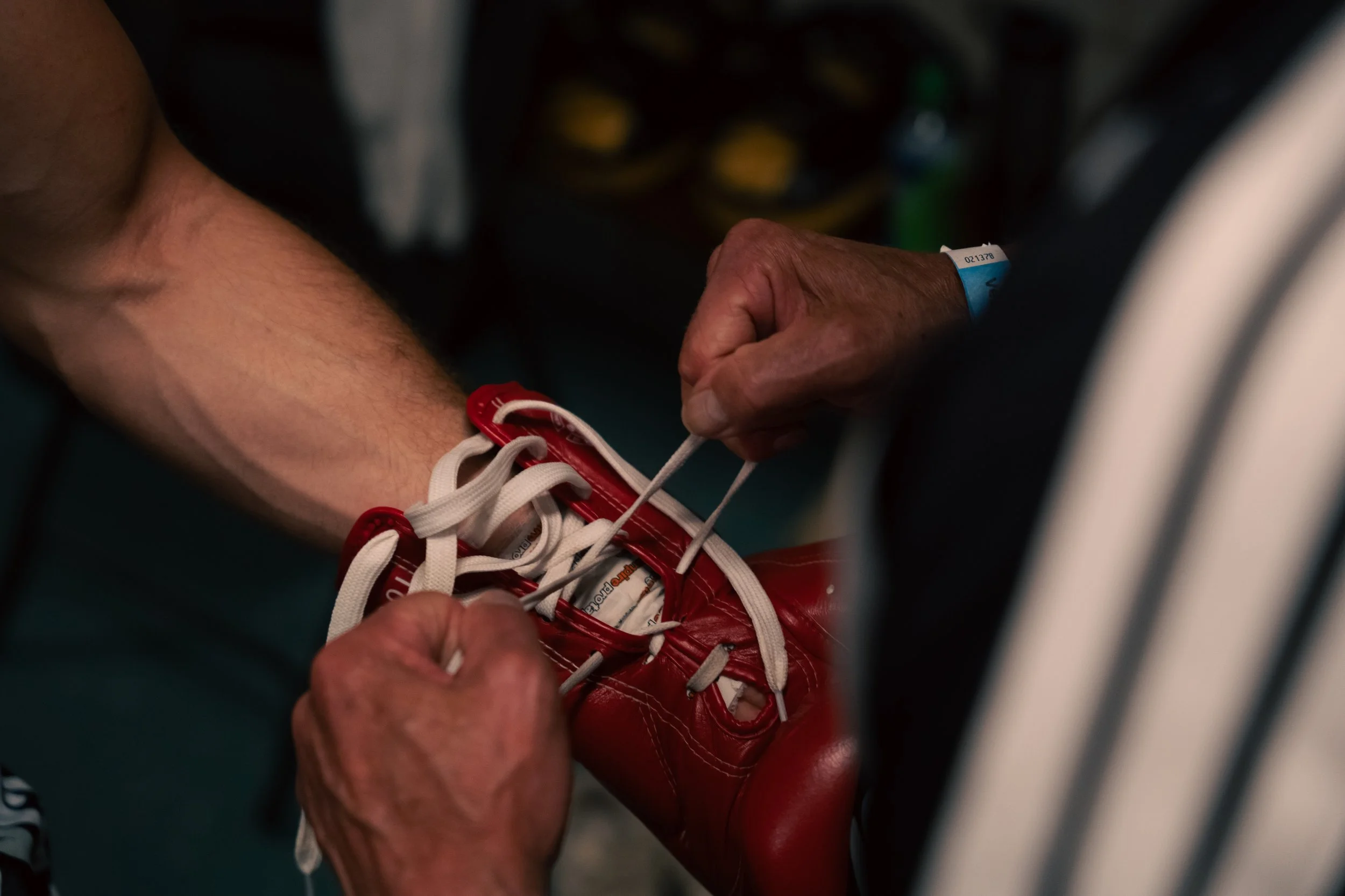 A close-up of a person's hand tying shoelaces on a red athletic shoe. The person is wearing a wristband. The background is blurred.