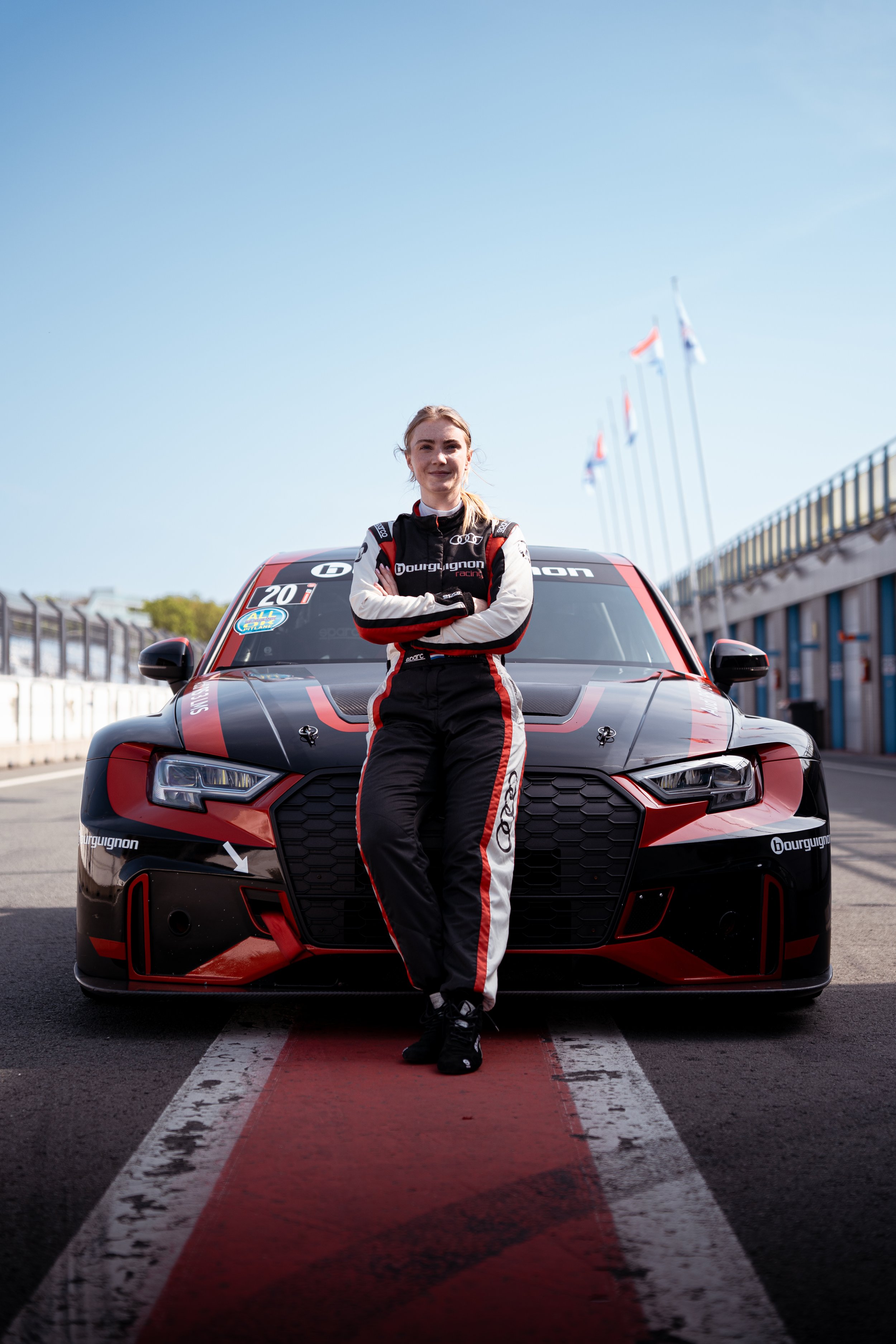 A female race car driver in a racing suit standing in front of a red and black race car on a race track, with her arms crossed.