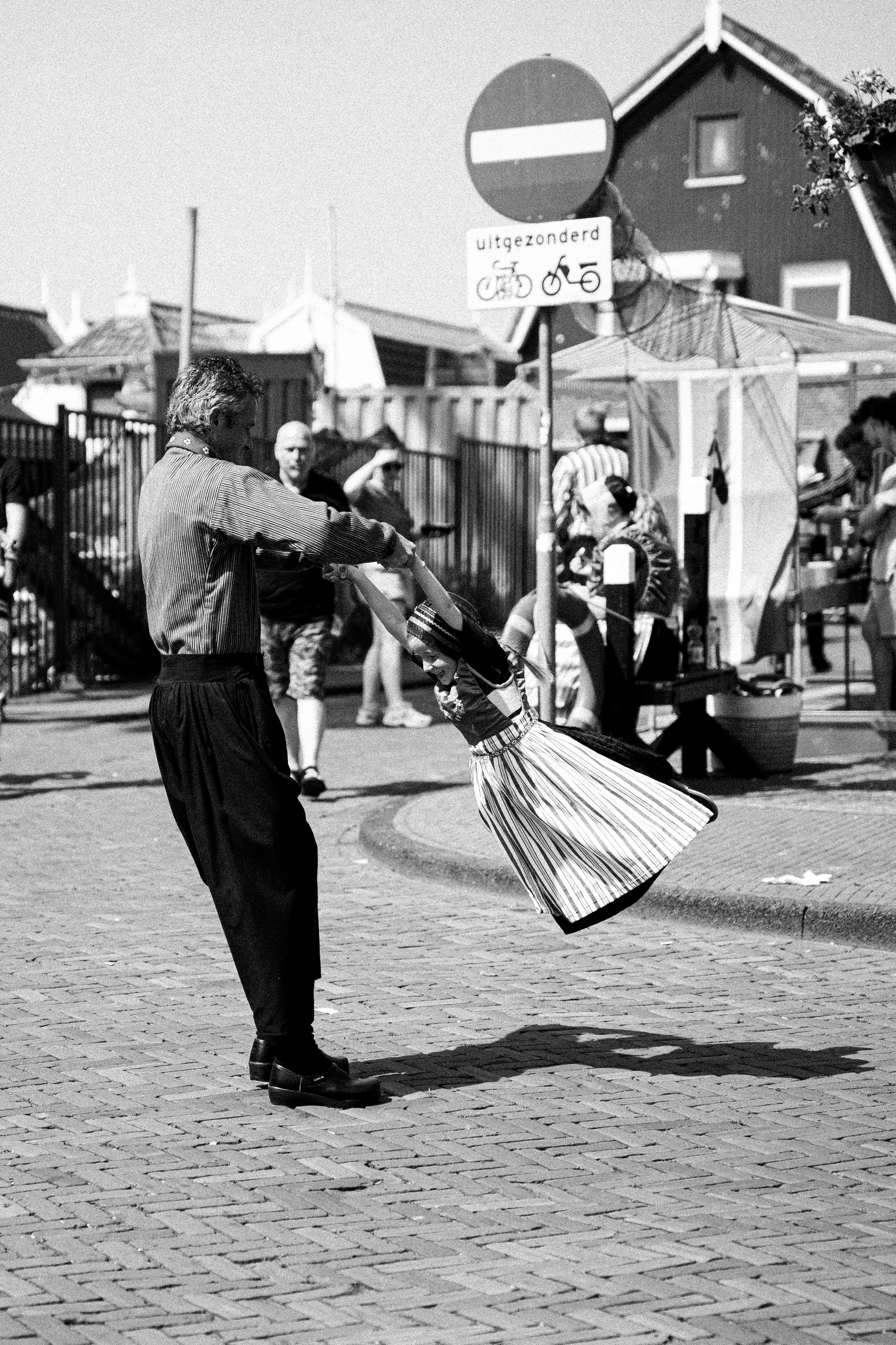 A black and white photo of a man and a woman dancing outdoors on a paved area, with several people in the background and a house with a garden on the right side.