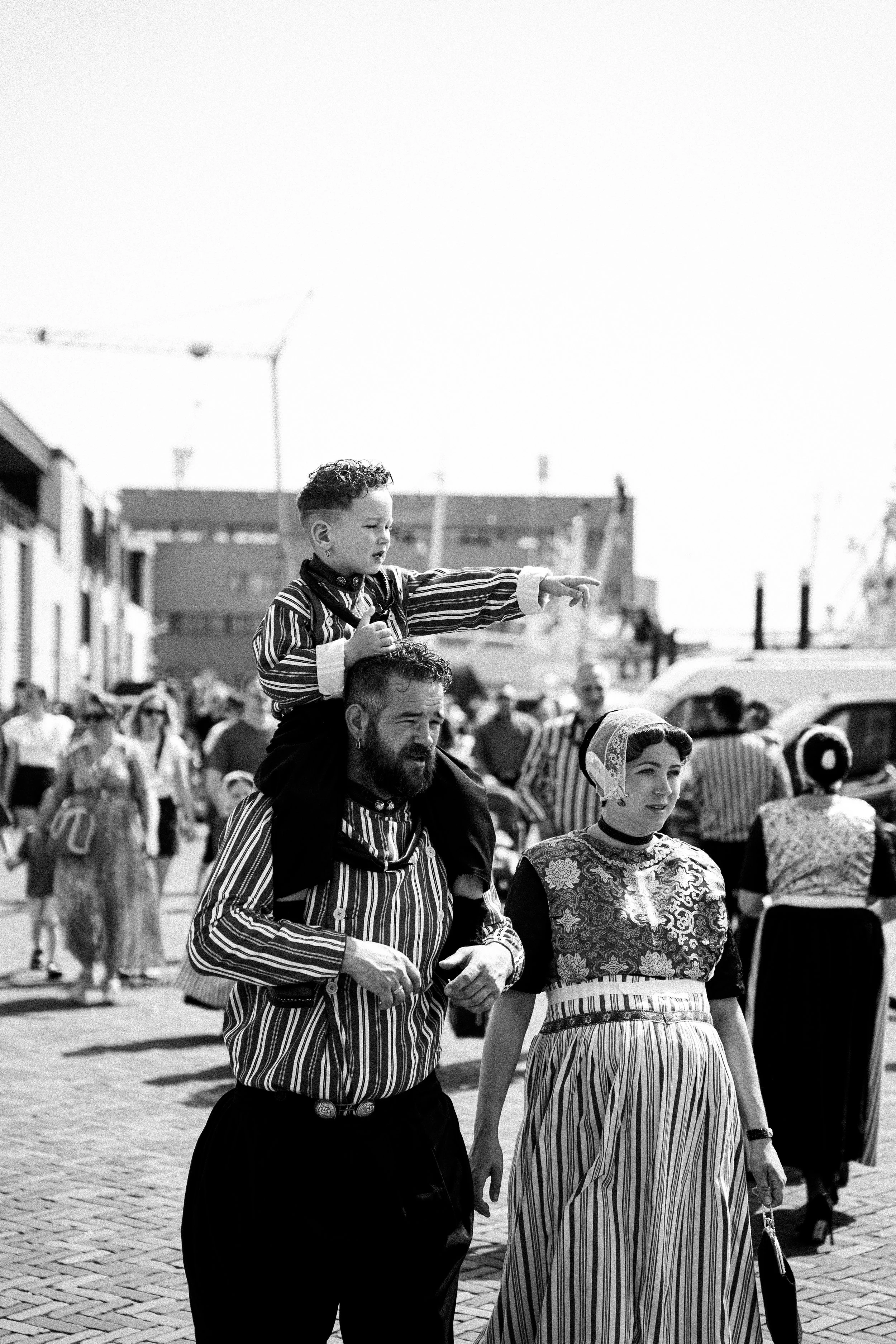 A black and white photo of a crowd in a public square with buildings and lamp posts in the background. In the foreground, a man with a beard, wearing a striped shirt, carries a young boy with curly hair on his shoulders. The boy is pointing at someth