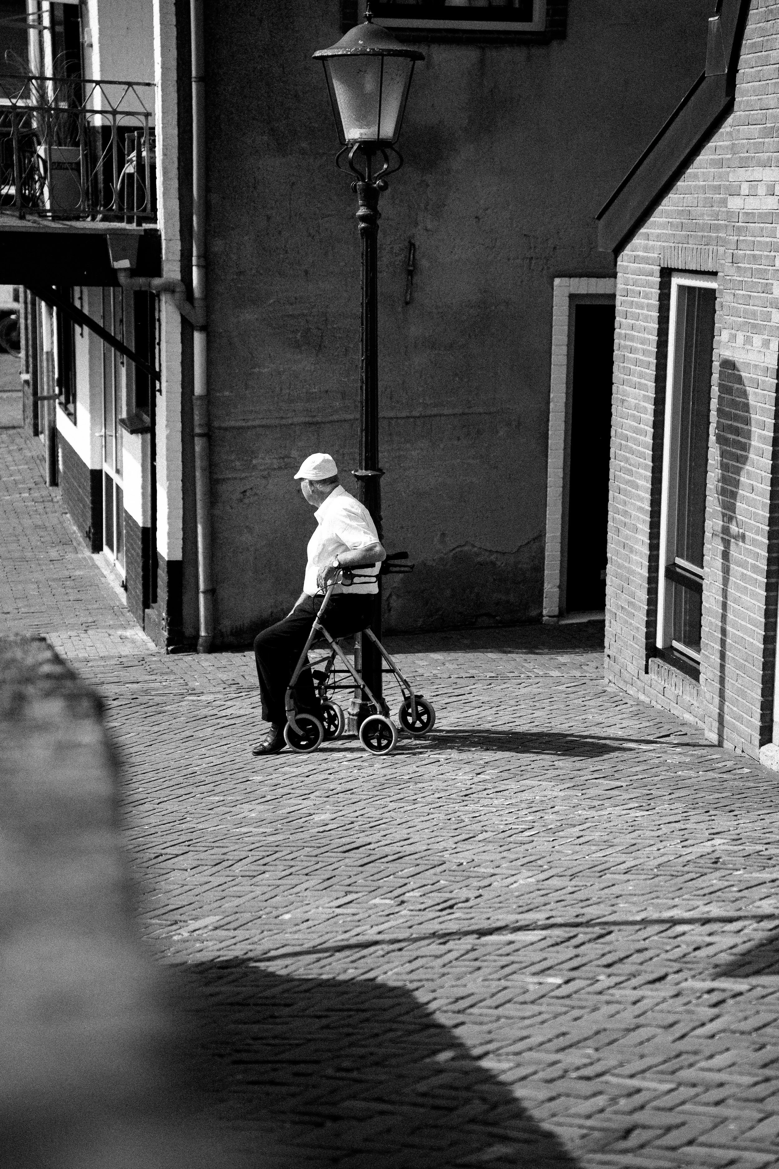 A man sitting on a chair with a walker beside him on a cobblestone street, with brick and stucco buildings and a streetlamp in the background.