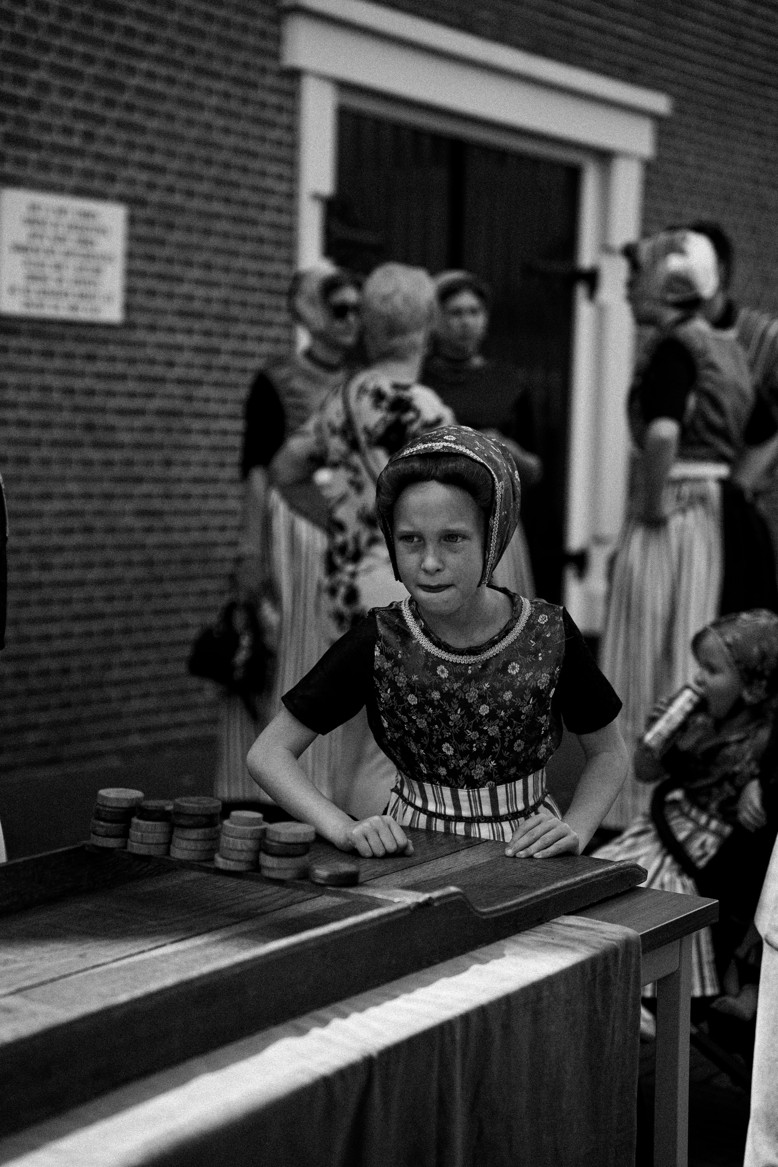 A young girl wearing a traditional dress and headscarf standing at a table with a chessboard, surrounded by children in ethnic costumes, possibly in a cultural performance or celebration.