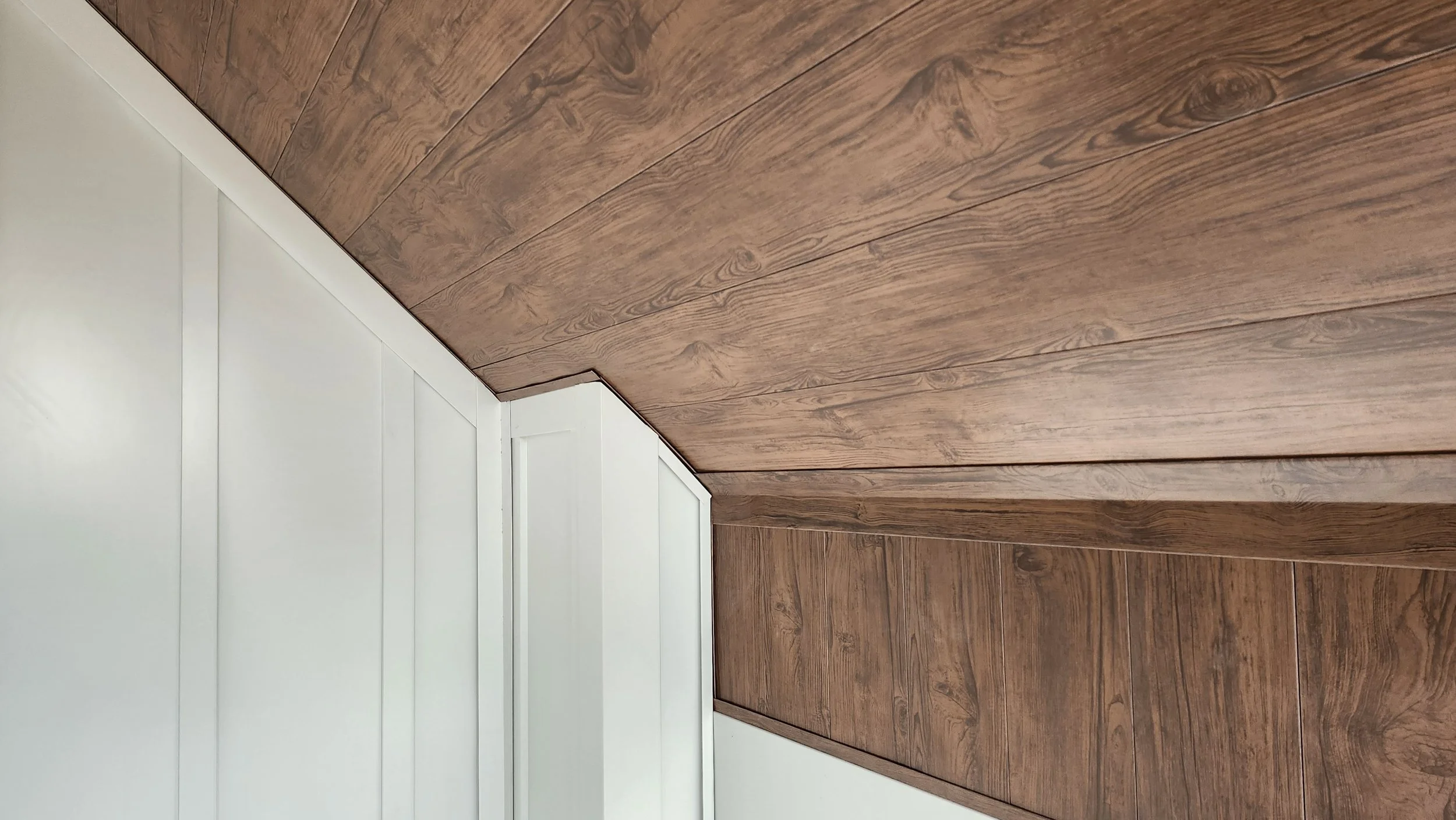 A corner of a room showing white paneling on the lower wall and brown wooden flooring and ceiling with visible wood grain.
