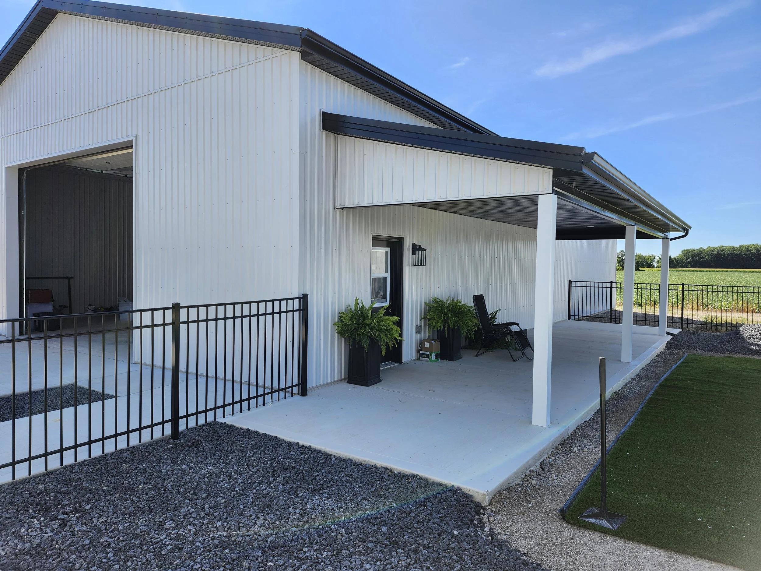 Back view of a white metal building with a covered porch, two black lawn chairs, green potted ferns, a small window, and a black lantern, with an open garage door and a fenced yard, under a blue sky.