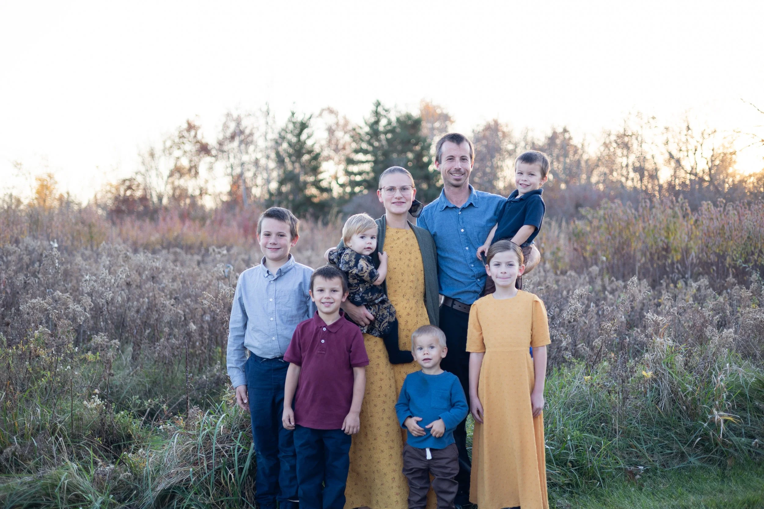 Family of nine posing outdoors in a field during sunset, with trees in the background