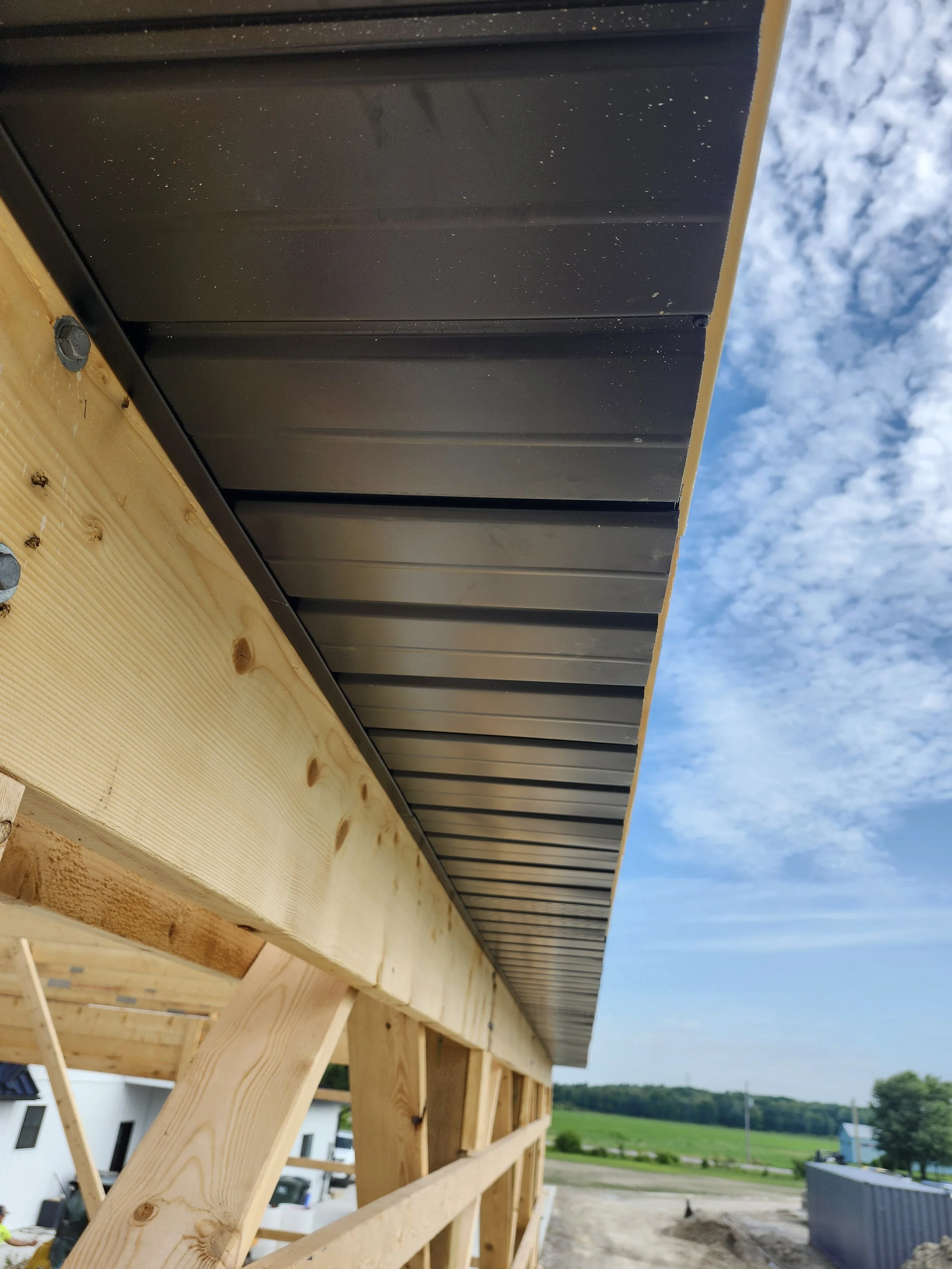 Construction site showing the underside of a building's roof with black metal siding, wooden framing, and an open outdoor view of blue sky and green fields.