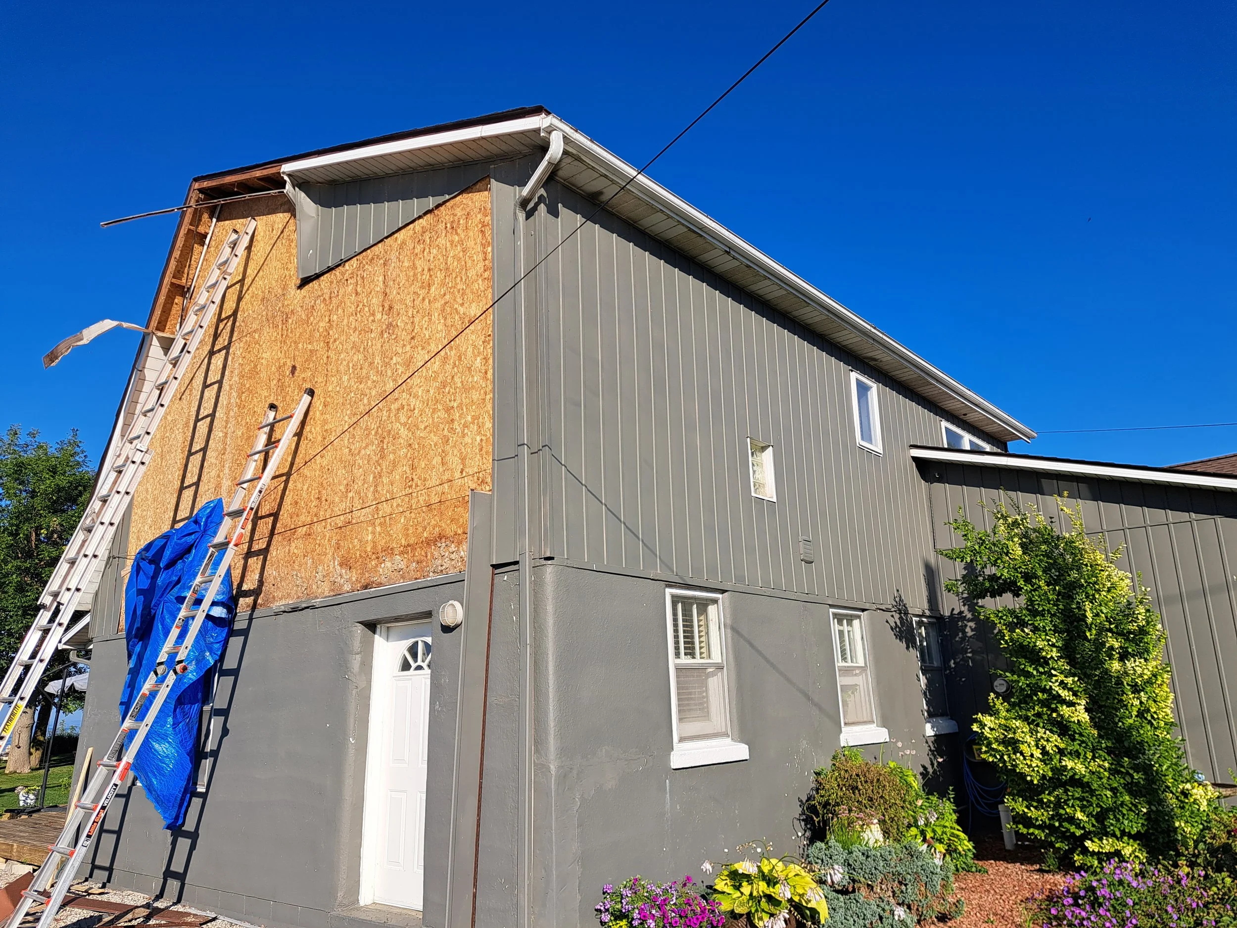 A house undergoing renovation with a section of the upper wall covered in plywood, ladders leaning against it, and a blue tarp hanging from one ladder. The house has gray siding, several windows, and a garden with plants and flowers in front.