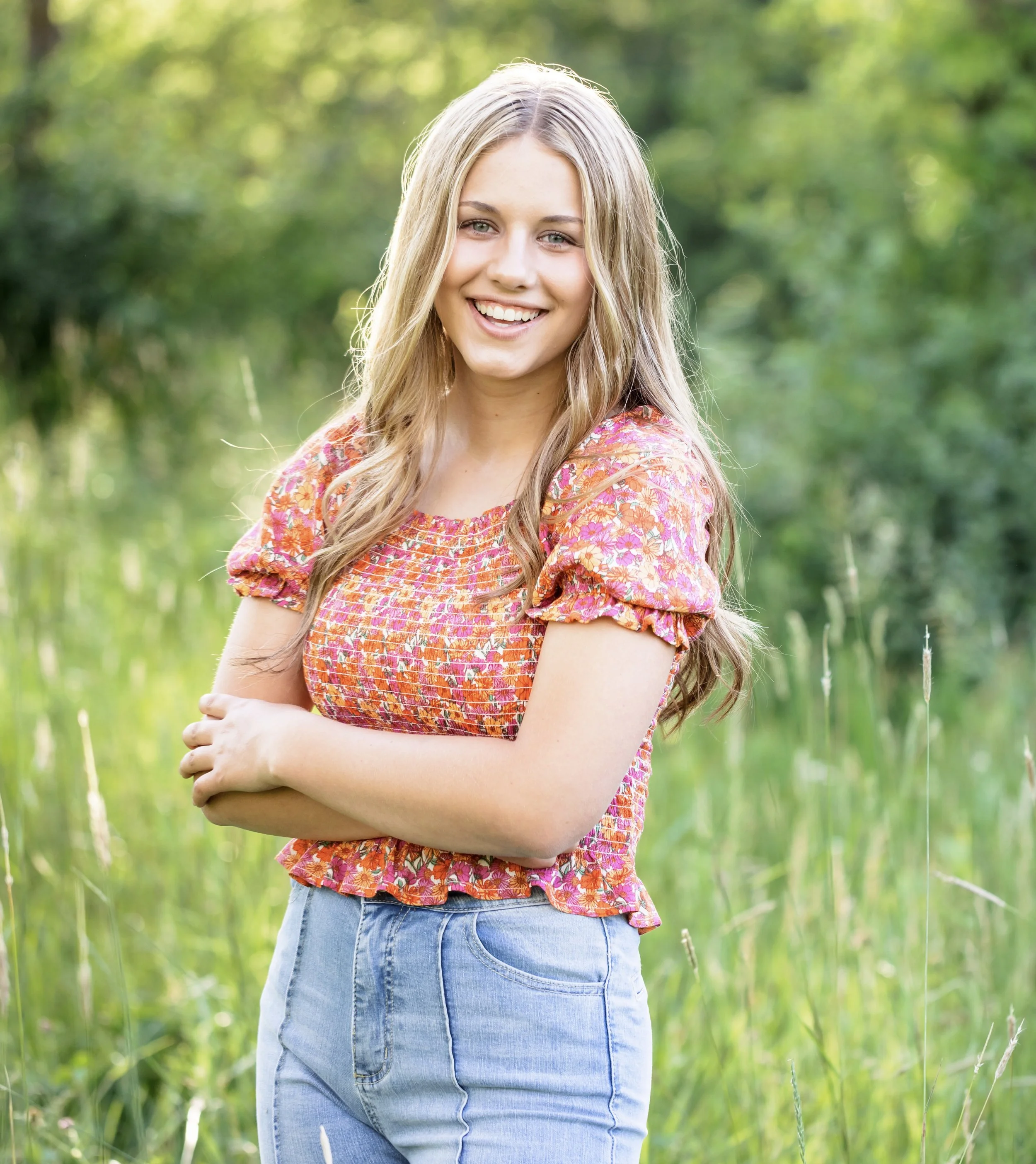 Young woman with long blonde hair smiling, wearing a pink floral top and blue jeans, standing outdoors in a green field.