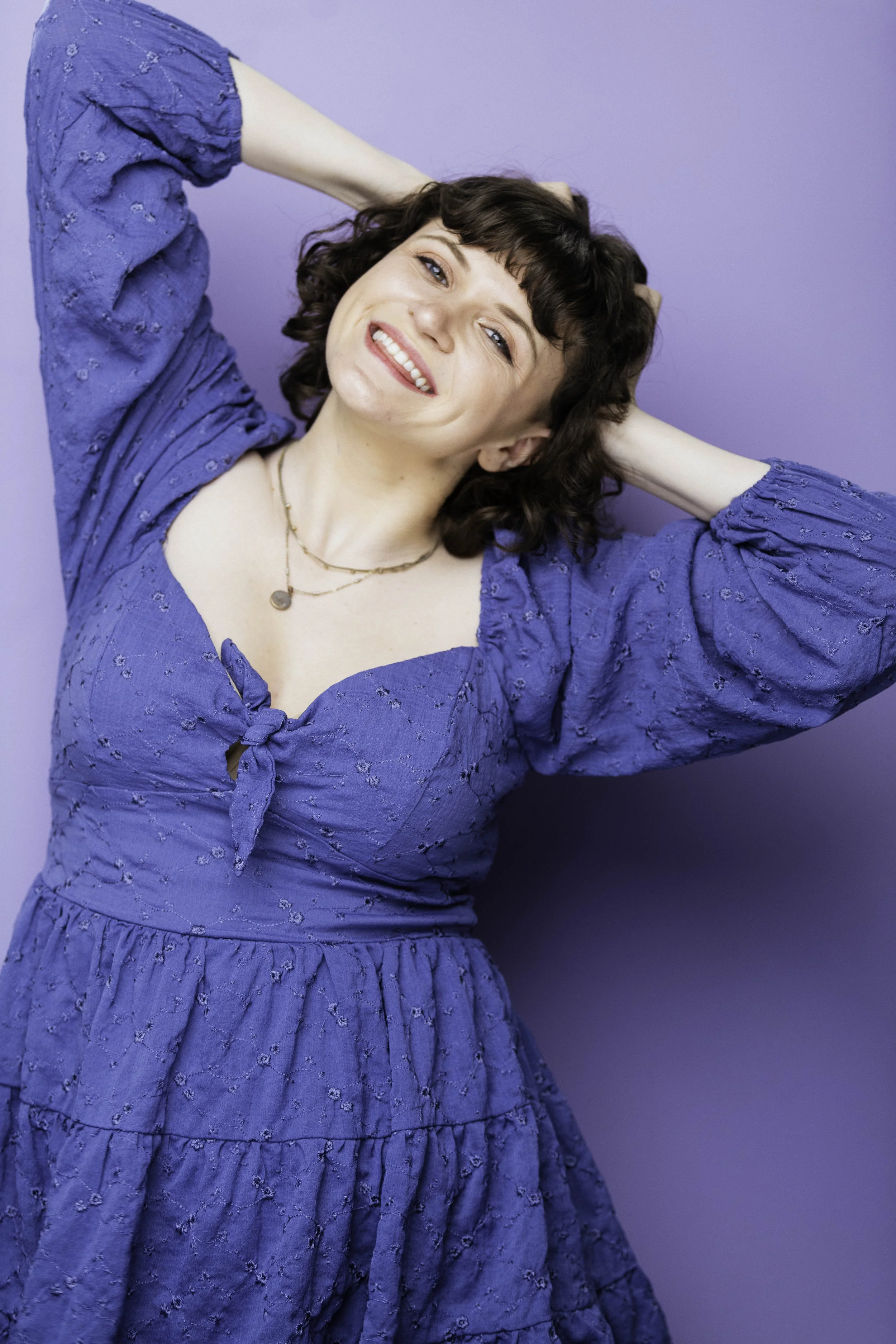 Julia Duffy, a young white woman with chin length curly brown hair and bangs. She has blue eyes and smiles at the camera playfully with a little bit of tongue poking between her teeth. Both her hands are in her hair, as if she is ruffling her curls.