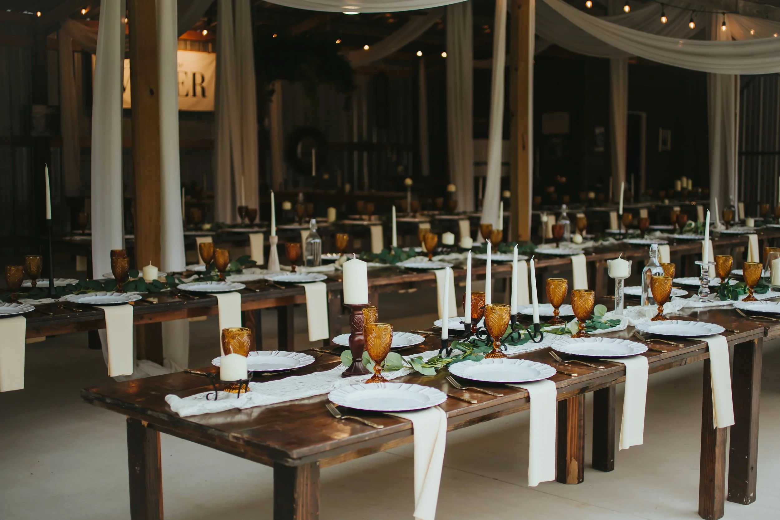 Long rustic wedding reception table decorated with white cloths, amber glasses, white candles, green foliage, and black candle holders in a barn style venue.