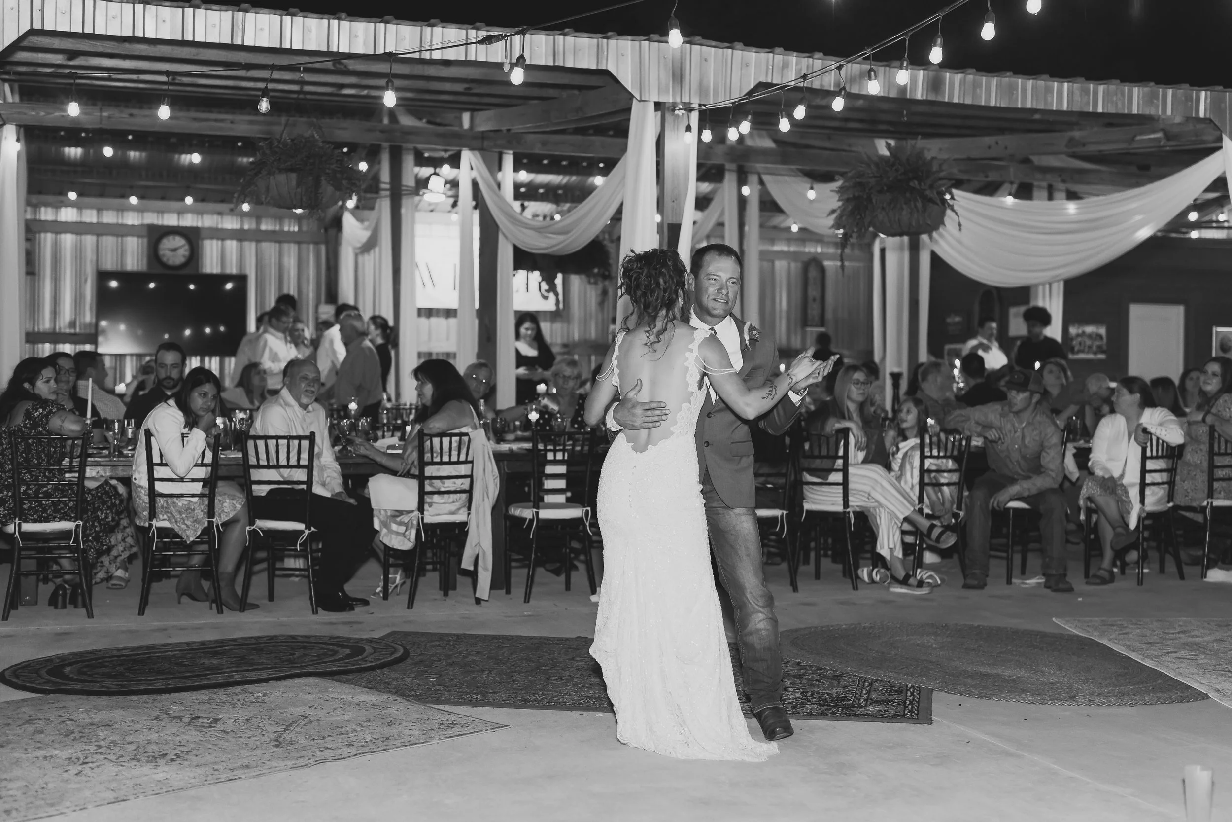 A bride and Father dancing at the wedding reception, with guests seated at tables in the background, decorated with drapes, hanging lights, and plants.