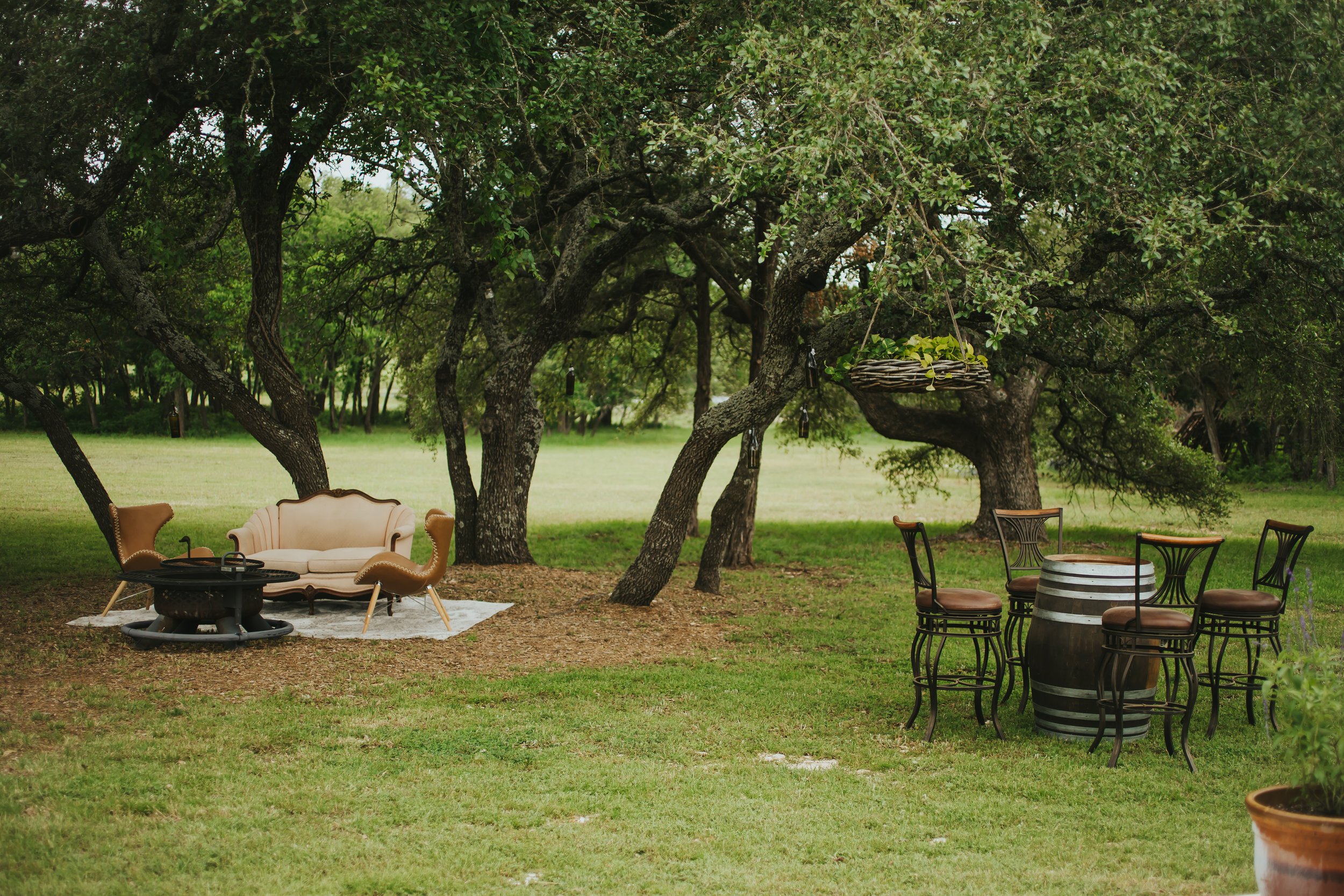 Outdoor patio area under large oak trees with vintage furniture including a cream-colored sofa, four chairs around a barrel table, and a hanging basket with plants.