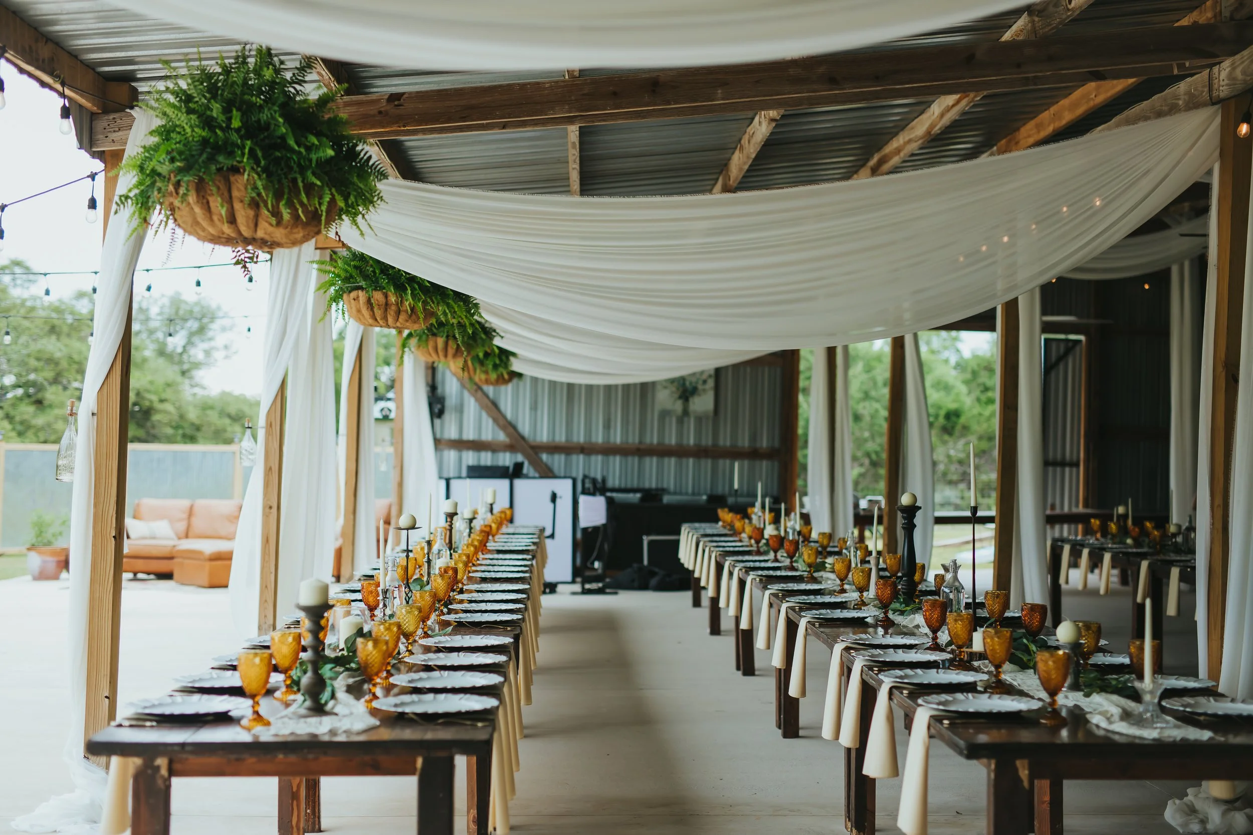 Long banquet tables set with plates, glasses, candles, and decorations in a rustic open-air venue with draped white fabric and hanging greenery.