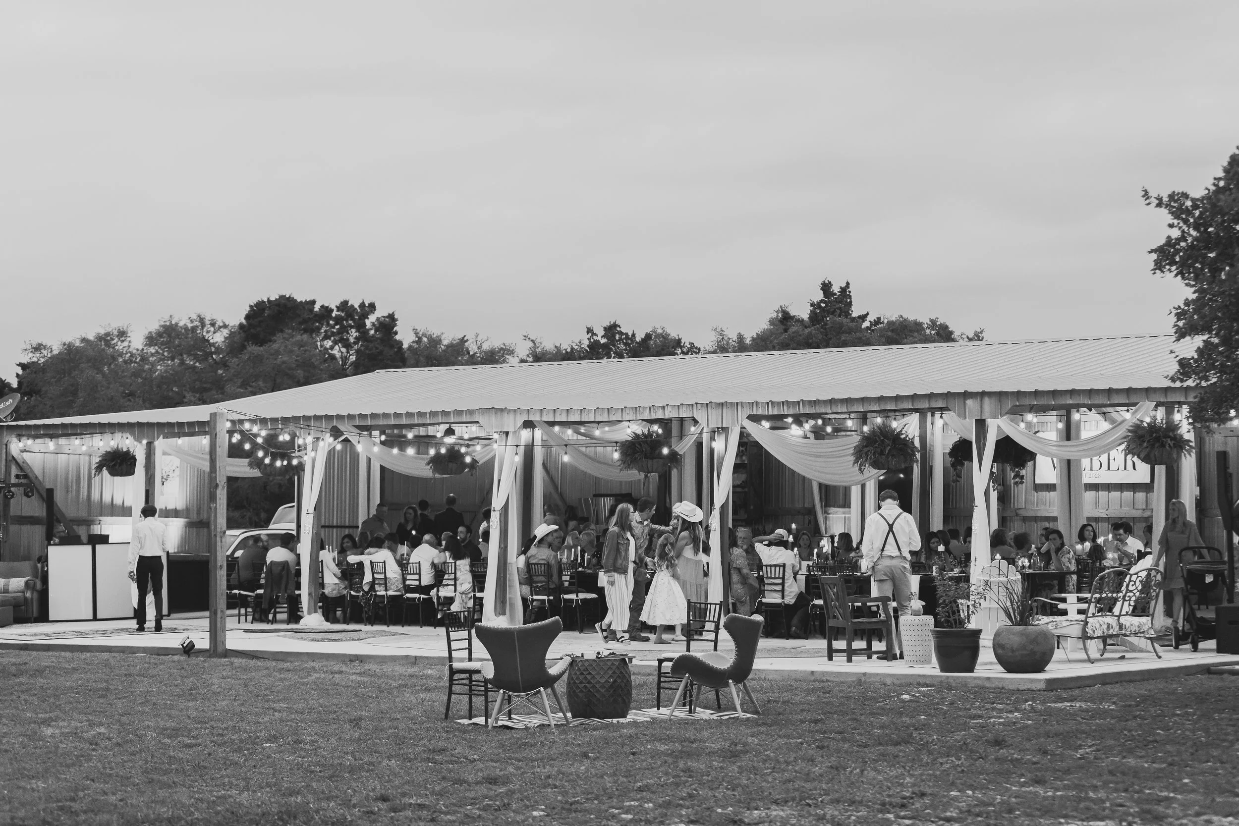 A black and white photo of an outdoor social event or wedding reception with string lights and hanging plants. People are seated at tables inside and some are standing and mingling outside the tent. The scene is set in a grassy area with outdoor chairs and tables in the foreground.