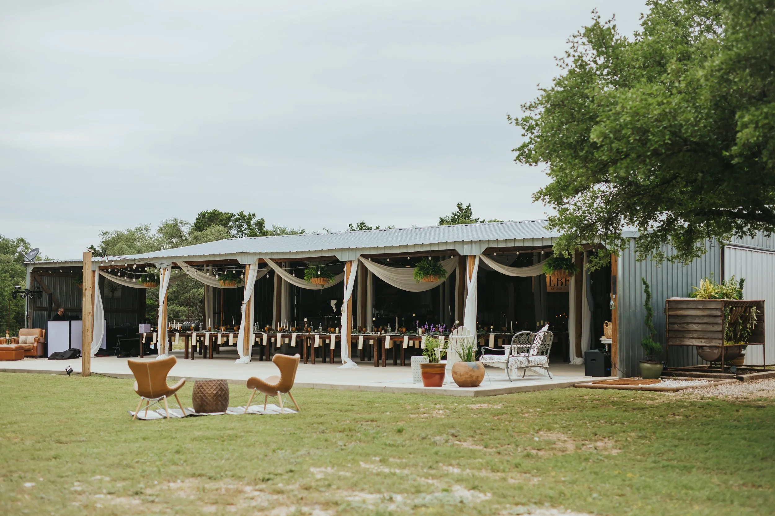 An outdoor event space with a long table decorated with white drapery and greenery, set inside a metal building. There are additional seating areas with armchairs and potted plants on the grass.