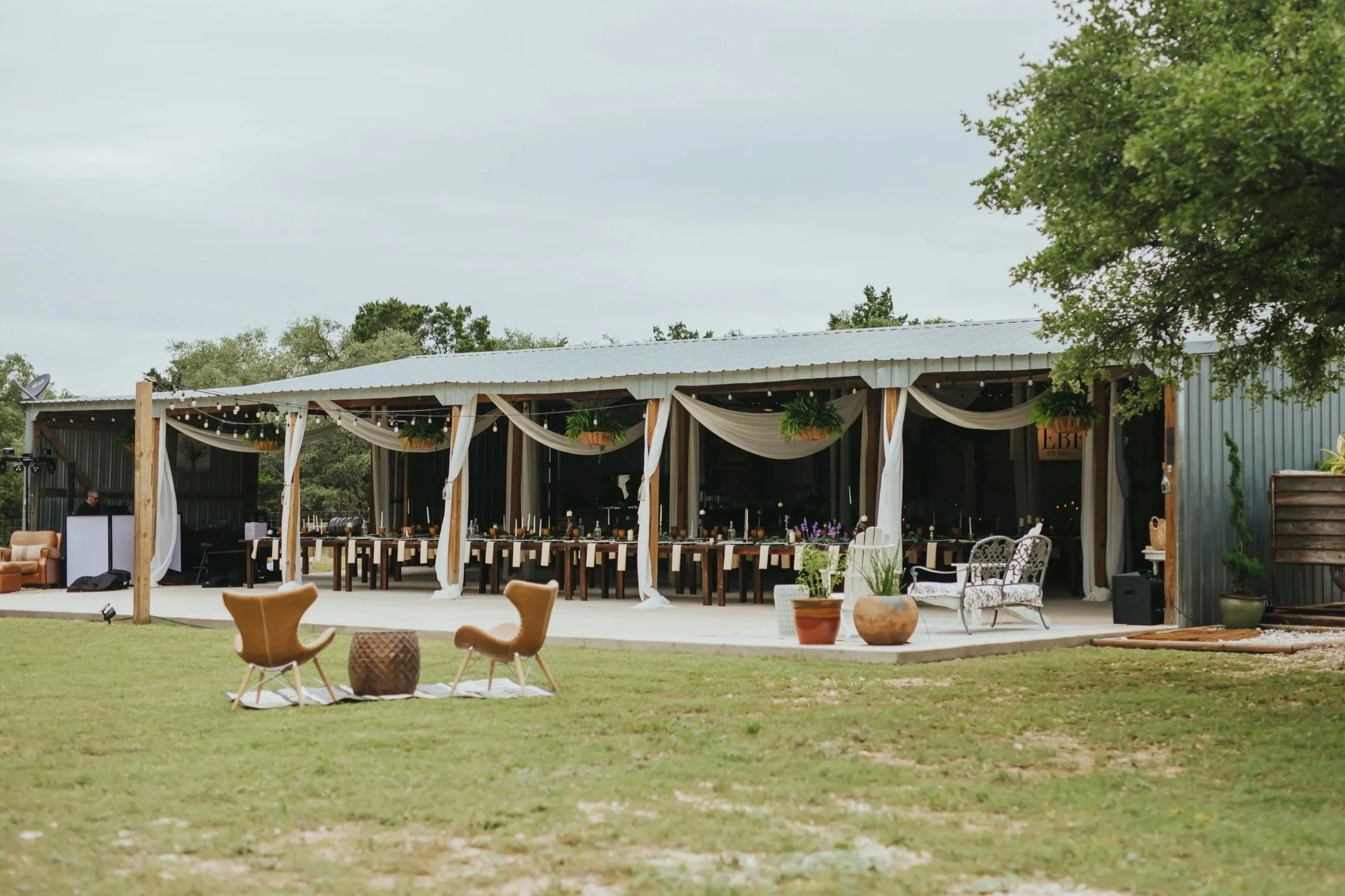 Outdoor wedding or event space, decorated with fabric swags, hanging plants, and string lights, surrounded by greenery and outdoor furniture.