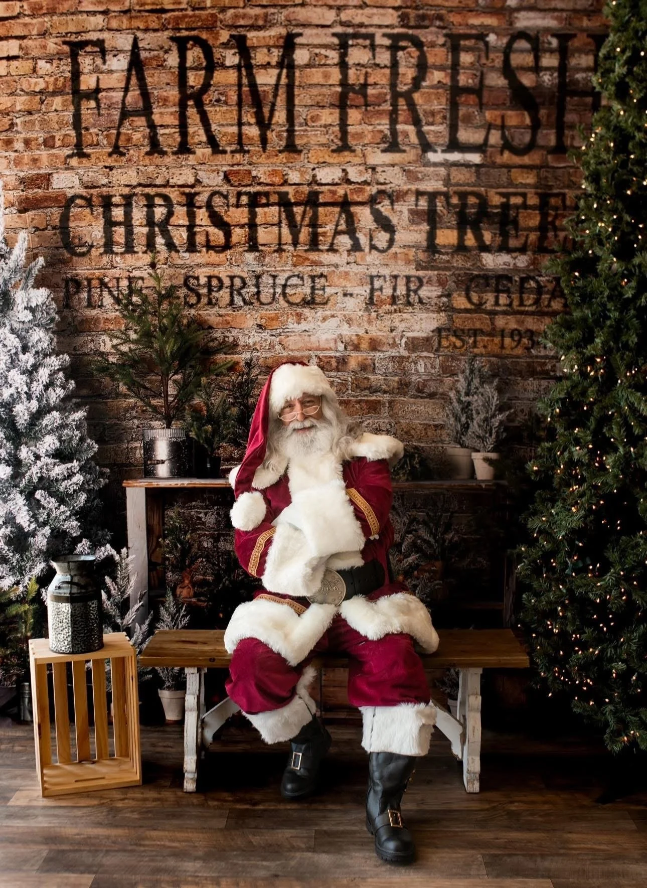 Santa Claus sitting on a wooden bench with a brick wall, Christmas trees, and holiday decorations in the background.