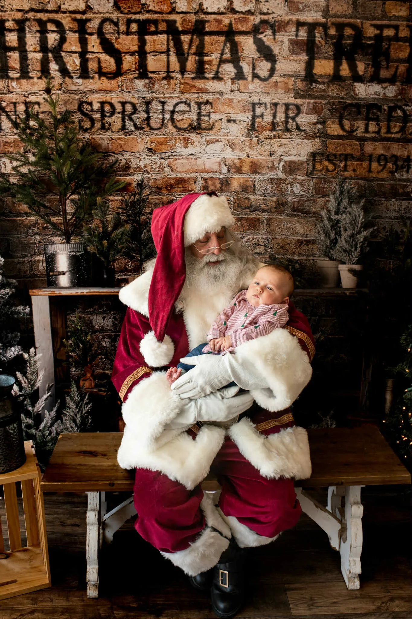 Santa Claus, dressed in a traditional red suit with white fur trim, holding a young child in his arms. The setting has a rustic brick wall backdrop with Christmas trees and holiday decorations.