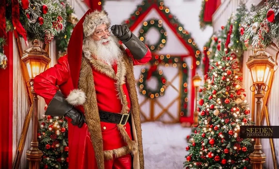 Santa Claus in a red outfit with a fur-trimmed cape, standing inside a decorated holiday scene with Christmas trees, wreaths, and warm lighting.