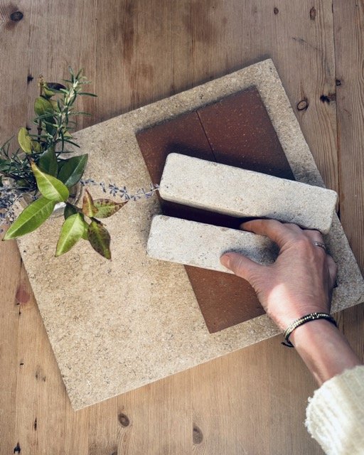 A person's hand is placing a brick onto a surface with various tiles and a potted plant nearby.