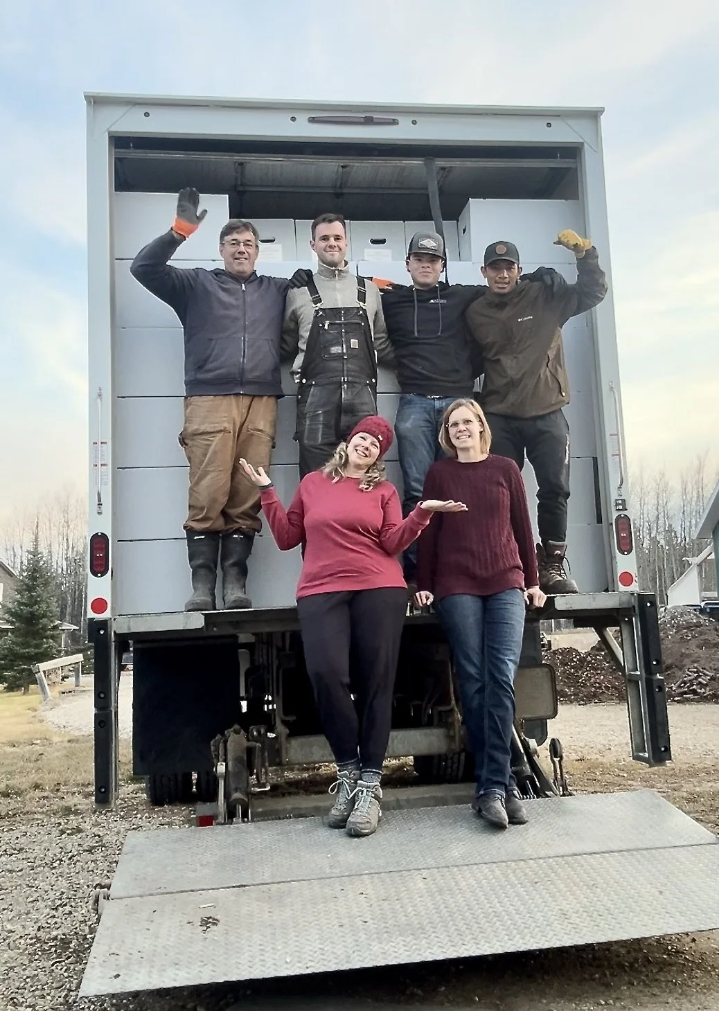 Group of five people standing on the back of a truck, with two women standing in front of the truck. One woman is wearing a red sweater and red beanie, the other a maroon sweater. The men are on the truck platform, some wearing work clothes and gloves, smiling and waving.