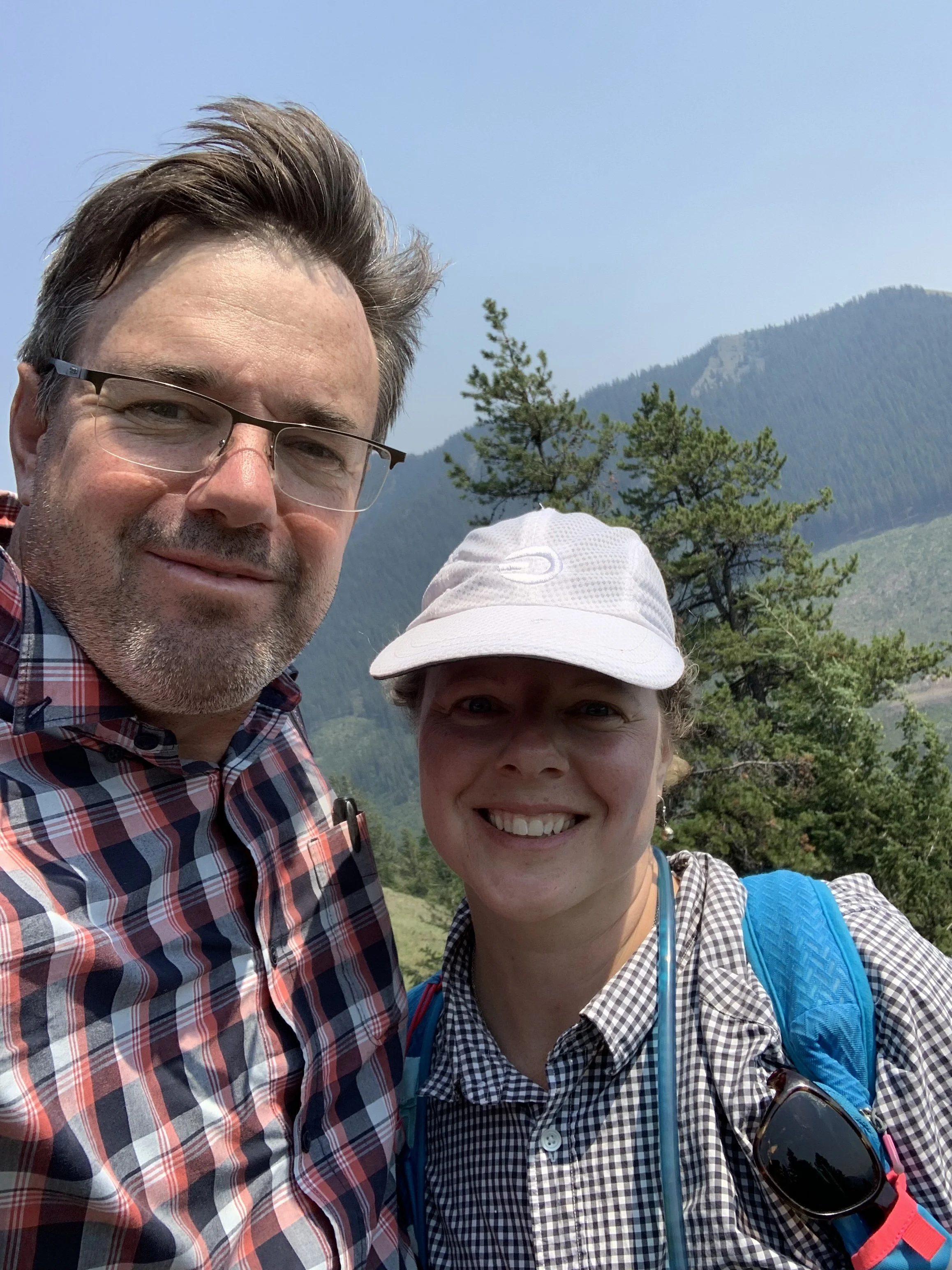 A man and a woman taking a selfie outdoors with mountains and trees in the background, smiling and dressed in outdoor clothing.