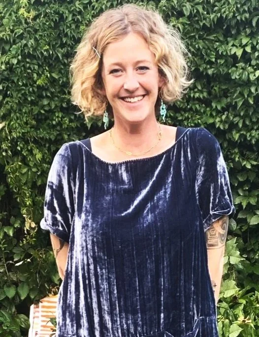 A woman with short, curly blonde hair and earrings, smiling in front of a green leafy background, wearing a dark blue velvet dress.