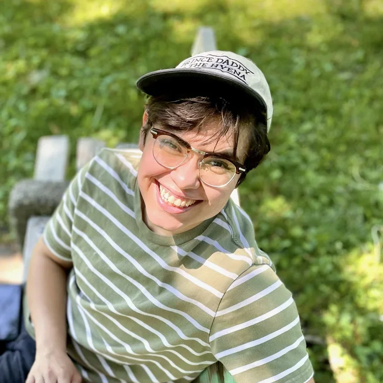 A young woman with short brown hair and glasses smiling, wearing a gray hat with text, and a green and white striped shirt, sitting outdoors on a park bench with green foliage in the background.