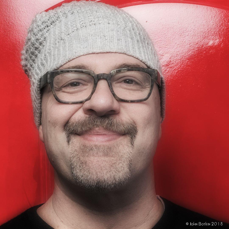 A man wearing glasses and a knit gray beanie hat smiling in front of a red background.