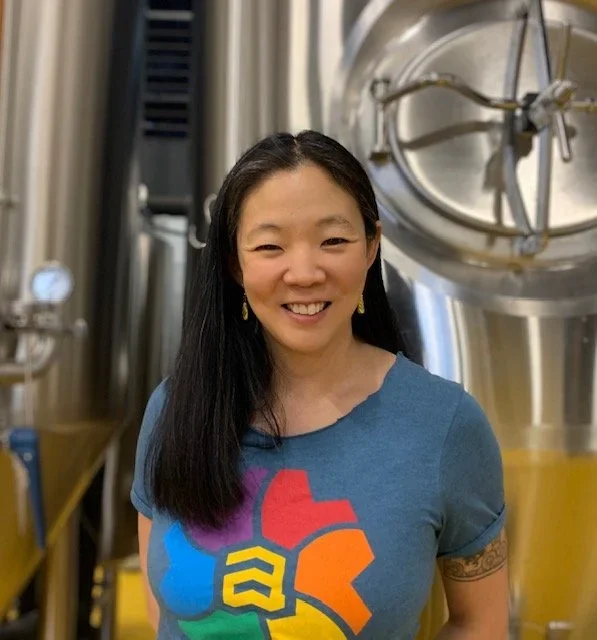 Woman with long dark hair smiling, wearing a blue t-shirt with a colorful abstract graphic, standing in a brewery or industrial kitchen with large metal tanks in the background.