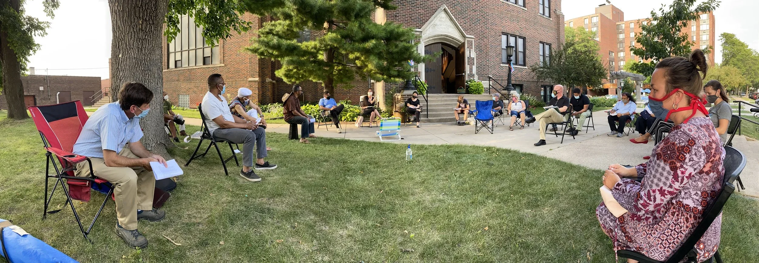 A group of people are sitting in a circle outdoors, socially distanced, wearing face masks, and participating in a gathering by a brick building with stairs, trees, and neighboring buildings in the background.