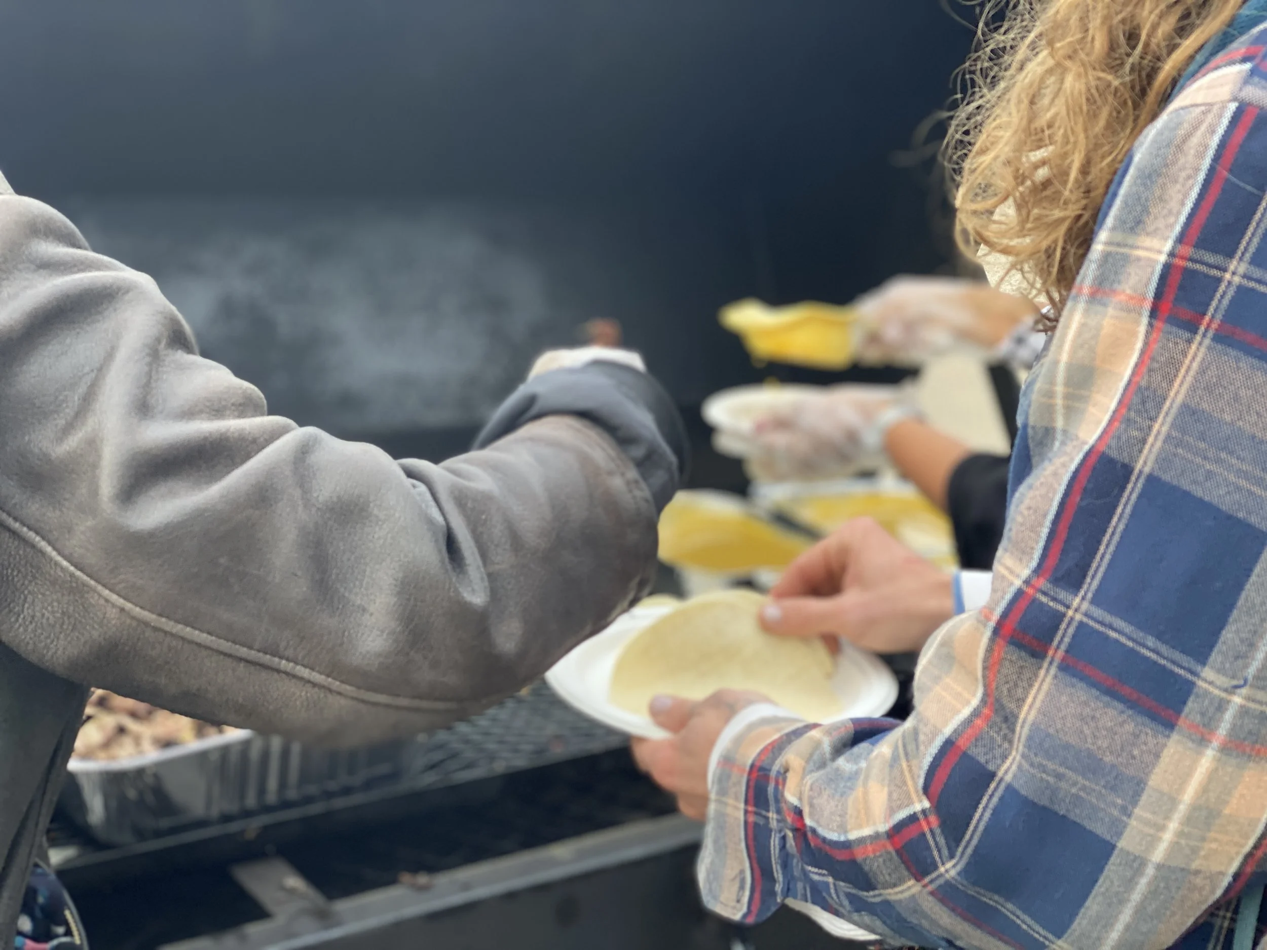 People wearing gloves serve taco shells onto plates at an outdoor cookout or barbecue.
