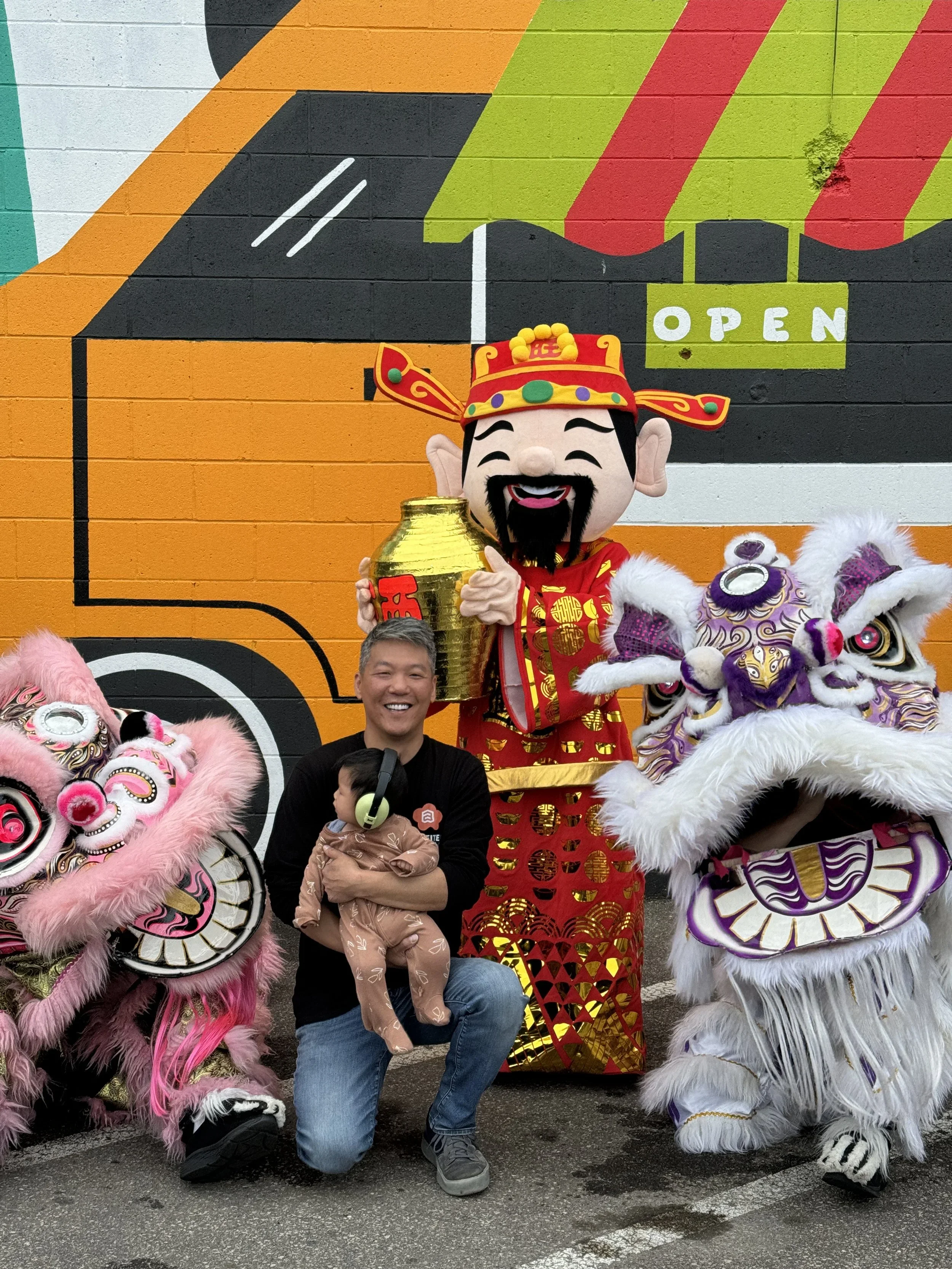 A man kneeling with a young child, surrounded by traditional Chinese lion dance costumes and a large cartoon figure in a Chinese outfit holding a gold jar, in front of a colorful wall with an "Open" sign.