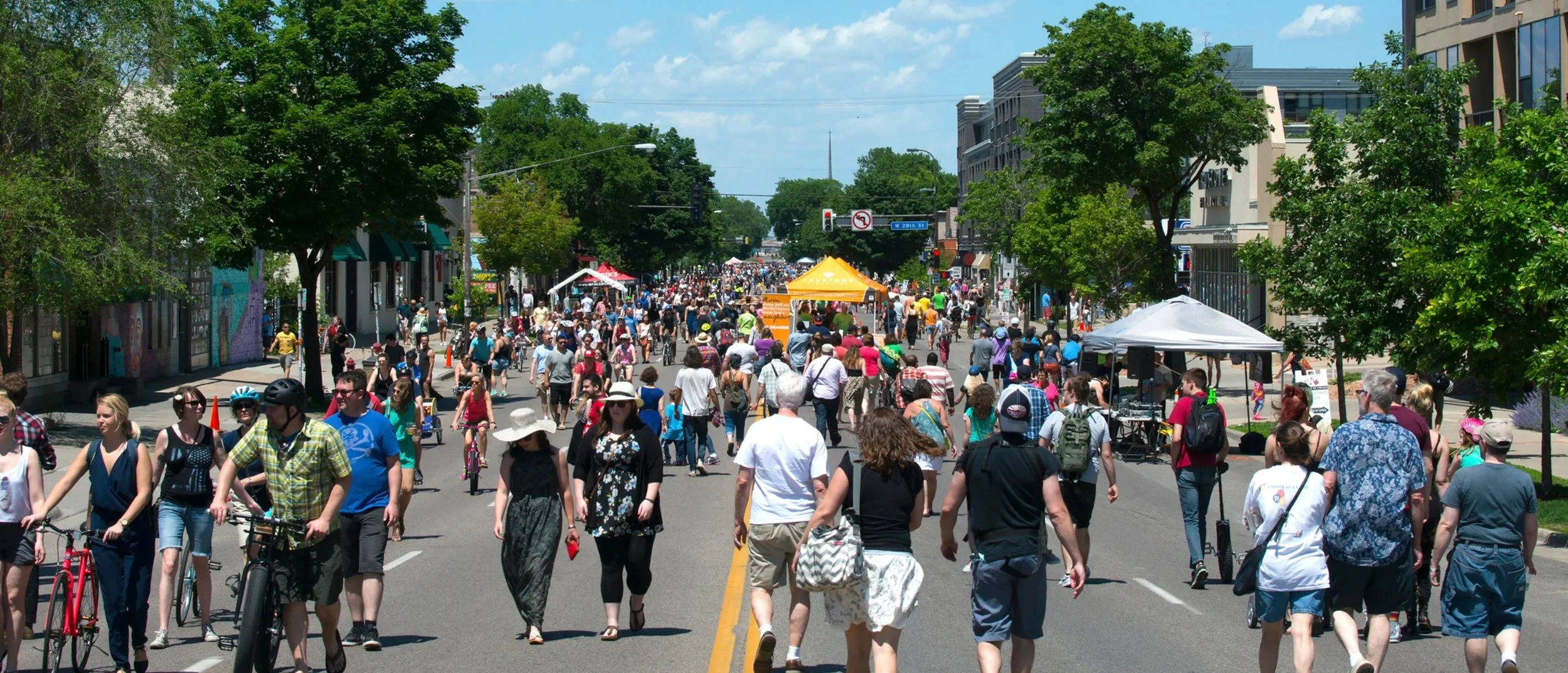 Crowd of people walking on a street during an outdoor event or festival, with tents and trees lining both sides of the street under a partly cloudy sky.