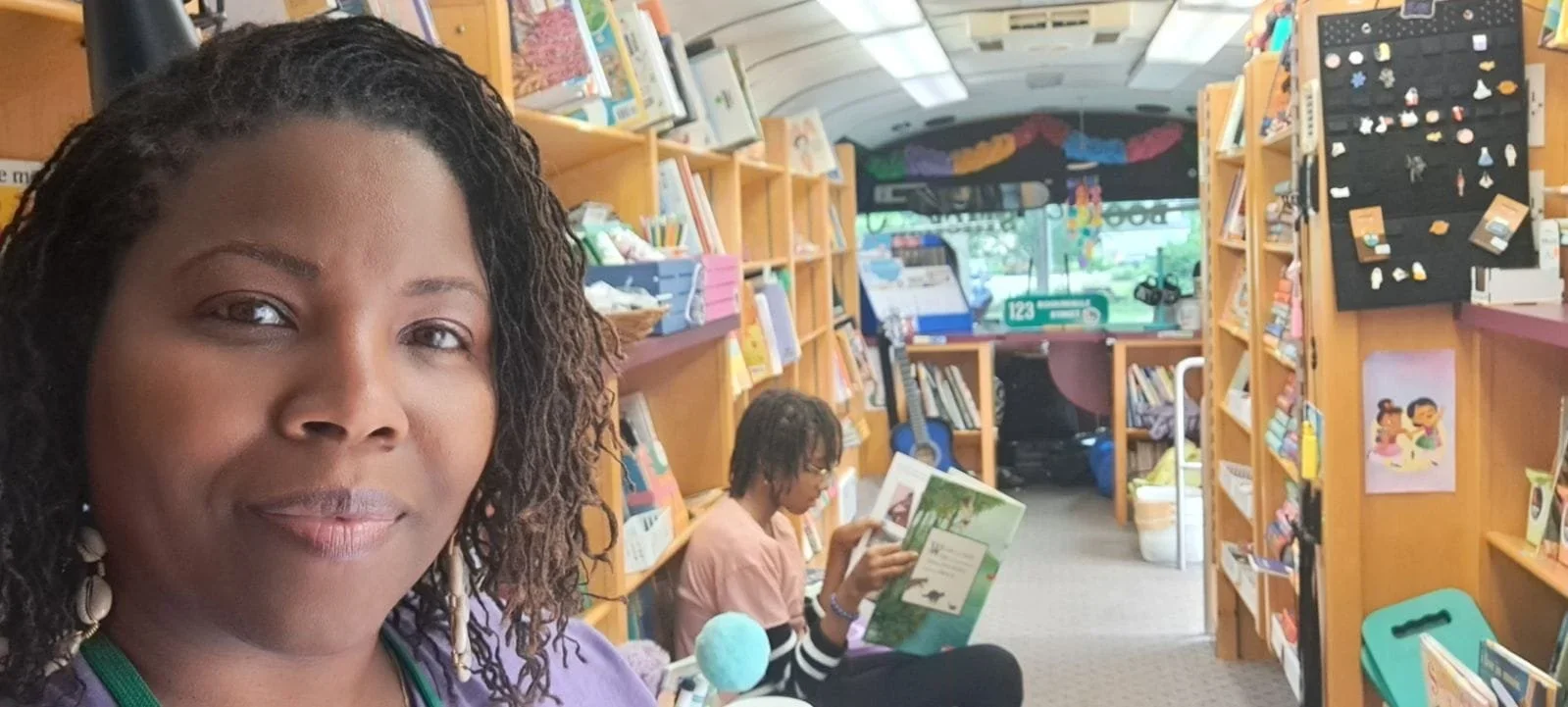 A woman with braided hair in front of a bookstore or library. Behind her, another person is reading a book among shelves filled with books, and the store has a colorful, inviting interior with posters and decorations.