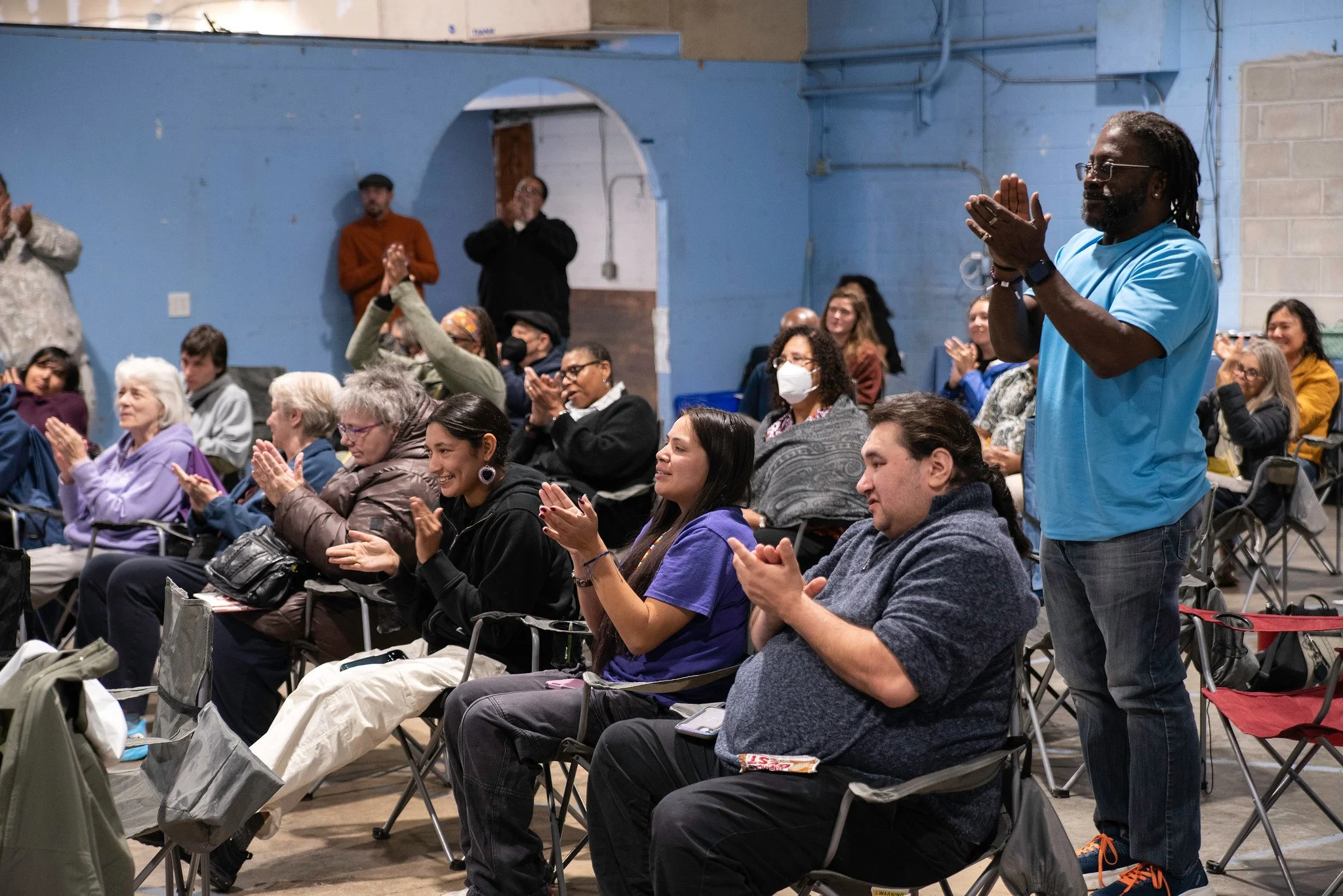 Group of diverse people attending an indoor event, some seated and some standing, applauding and showing appreciation.